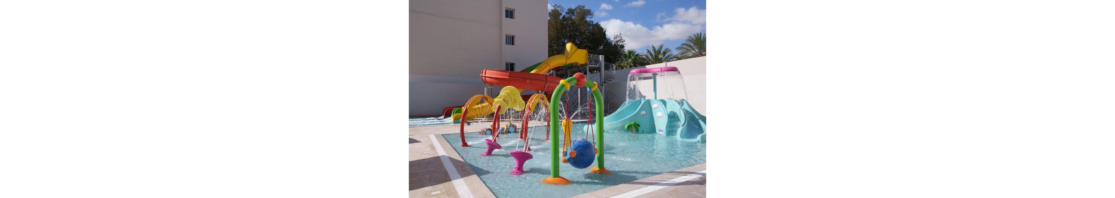 A vibrant children’s splash park features multiple interactive water structures in a shallow pool. Curved slides, spraying arches, and playful animal shapes create a fun and engaging environment. Bright colors like yellow, green, pink, and blue stand out against the neutral pool deck and surrounding buildings. Palm trees and clear skies in the background suggest a warm, sunny resort setting.