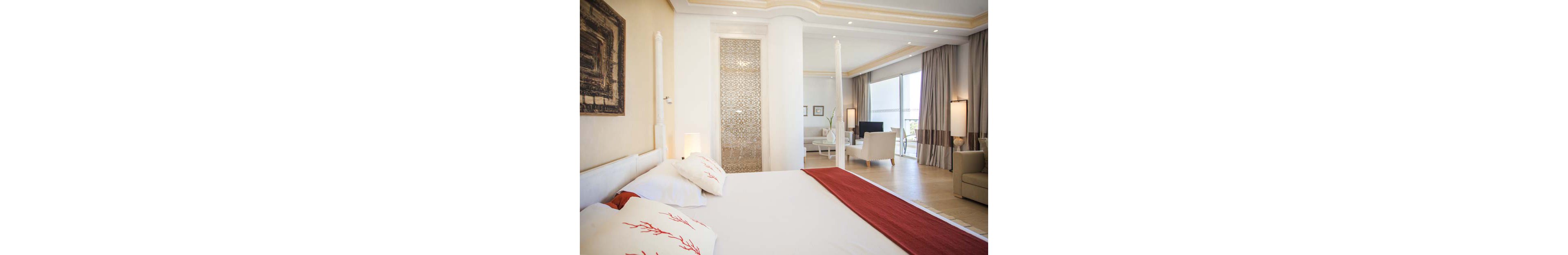 A spacious, modern hotel bedroom features a large white bed with red accents and decorative coral-pattern pillows. The room opens into a sitting area with white armchairs and a small table near floor-to-ceiling windows. Neutral beige walls, light wood flooring, and soft curtains create an airy, coastal atmosphere. Warm lighting and minimalist decor contribute to a calm, inviting space ideal for relaxation.