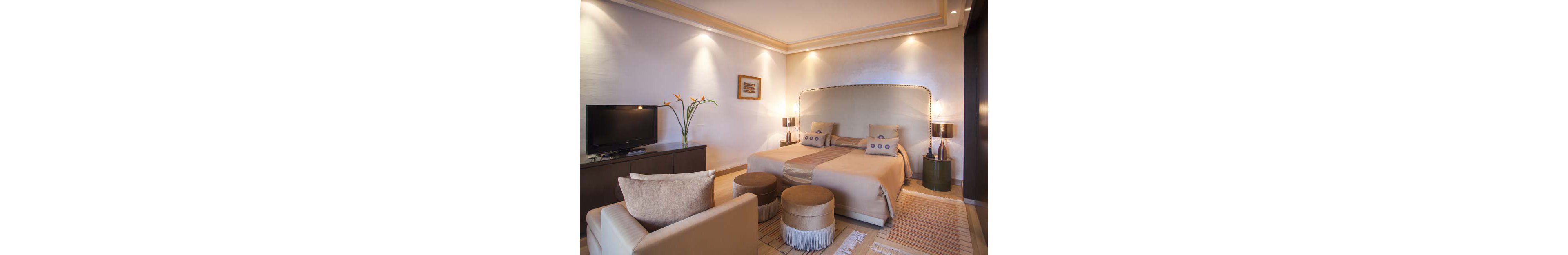 A contemporary hotel bedroom features a large double bed with coordinated beige bedding and accent pillows. The room includes a flat-screen television on a dark console, a sofa, and round upholstered stools at the foot of the bed. Soft wall lighting and a framed artwork create a warm, inviting atmosphere. Neutral tones, textured rugs, and a vase with flowers add subtle decorative details.