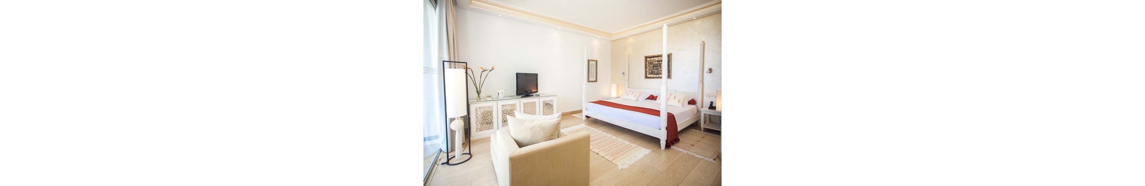 Spacious contemporary bedroom features a white four-poster bed with red runner and pillows. A cream armchair faces a cabinet holding a flat-screen television, kettle, and decorative vase. Light wood flooring, neutral walls, and soft lighting create an airy, minimalist atmosphere. Large windows with sheer curtains allow natural light to fill the room, highlighting the simple artwork and subtle decor.