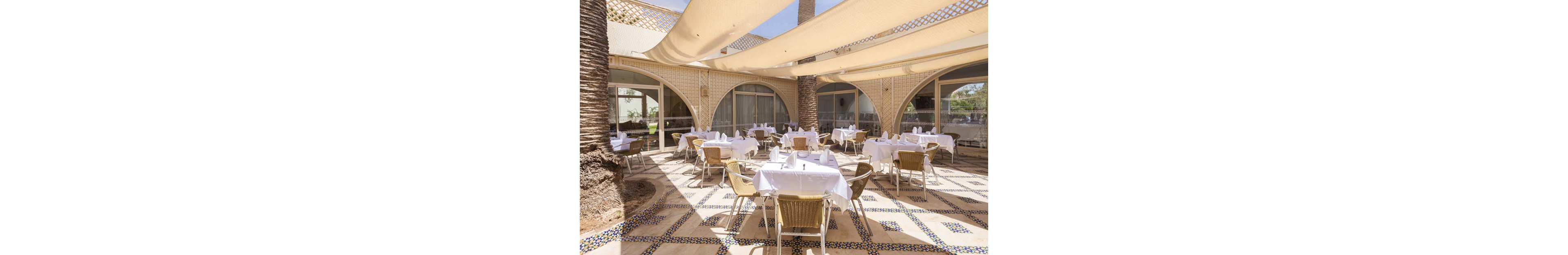 An elegant outdoor dining terrace is set with round tables covered in white tablecloths and neatly arranged place settings. Wicker chairs surround each table, creating a relaxed yet refined restaurant atmosphere. Large fabric canopies stretch overhead, casting soft shade across the tiled floor with intricate geometric patterns. Arched windows and palm trees frame the space, suggesting a warm, resort-like setting ideal for al fresco meals.