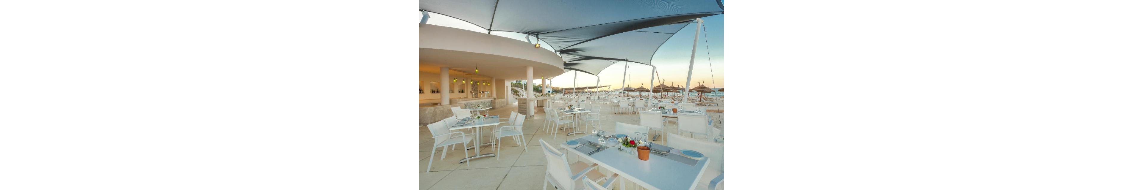 An open-air restaurant with white modern tables and chairs overlooks a sandy beach. The dining area is shaded by large fabric canopies attached to a curved, minimalist building. Place settings and small floral centerpieces are arranged neatly on the tables, ready for guests. In the background, straw parasols line the shoreline under a soft evening sky, creating a relaxed coastal atmosphere.