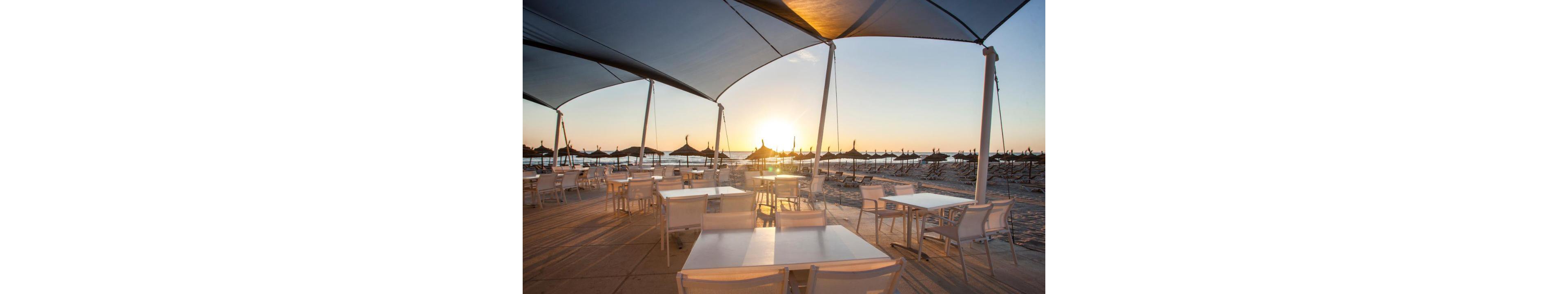 An open-air café terrace with white tables and chairs overlooks a sandy beach at sunset. Large shade canopies stretch overhead, casting soft shadows across the seating area. Straw parasols line the shoreline, silhouetted against the low sun over the calm sea. The warm golden light and clear sky create a tranquil, relaxing coastal atmosphere.
