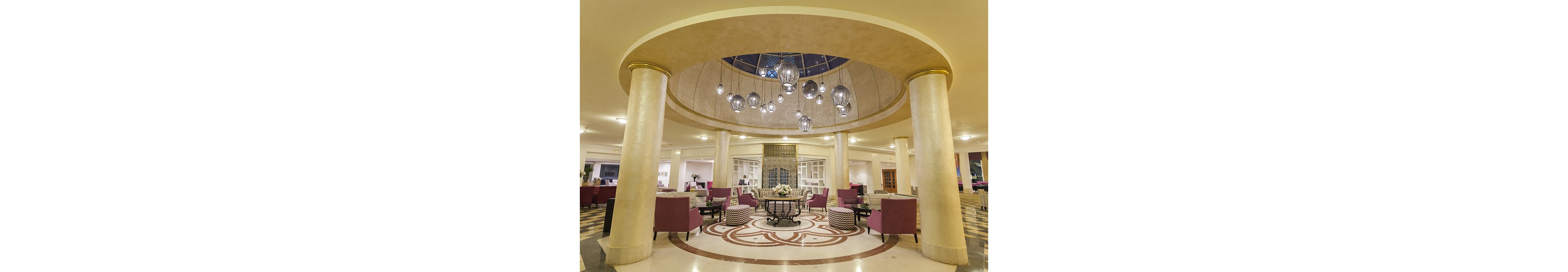 A spacious hotel lobby features a grand circular seating area framed by tall cream-colored columns. Contemporary pendant lights hang from a domed ceiling above a patterned floor medallion. Plush magenta armchairs and patterned ottomans surround a central table with floral arrangements. The background includes reception or lounge areas, creating a welcoming and luxurious atmosphere.