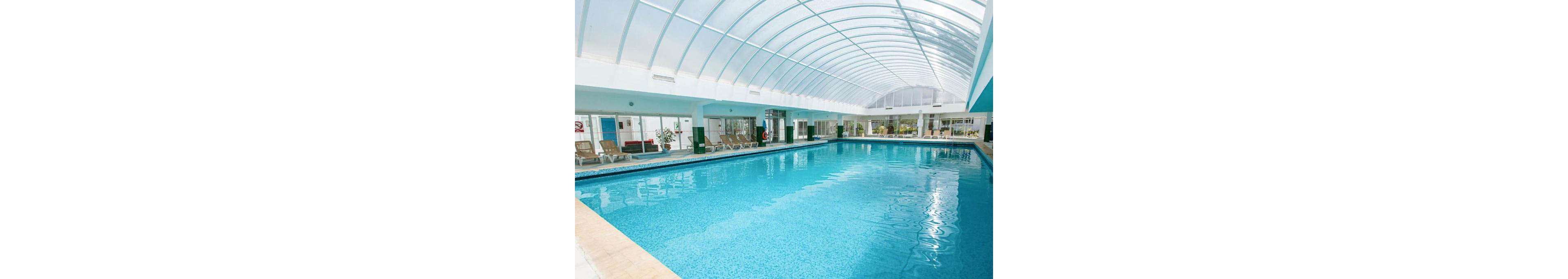 A large indoor swimming pool stretches beneath a high, translucent arched roof. Sunlight filters through the curved ceiling, illuminating the clear blue water and tiled pool deck. Lounge chairs line the sides of the pool, with a few potted plants and seating areas visible in the background. The clean, modern space suggests a hotel, resort, or wellness facility designed for relaxation and recreation.