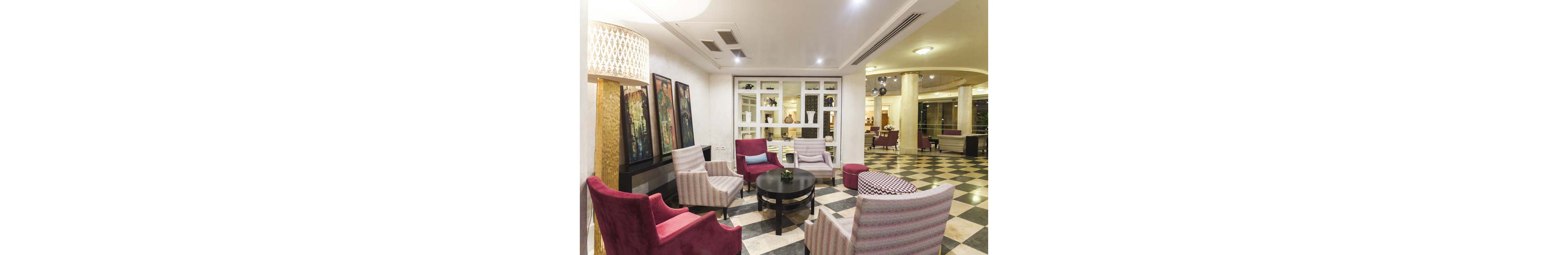 A contemporary hotel lobby lounge features a cozy seating area with upholstered armchairs arranged around a small round coffee table. The space is decorated with framed artwork on the wall and a large decorative lamp providing warm ambient light. In the background, glass shelving displays vases and decorative objects, while the checkered floor extends into the wider lobby. The overall atmosphere feels inviting and comfortable, suitable for guests to relax or socialize.