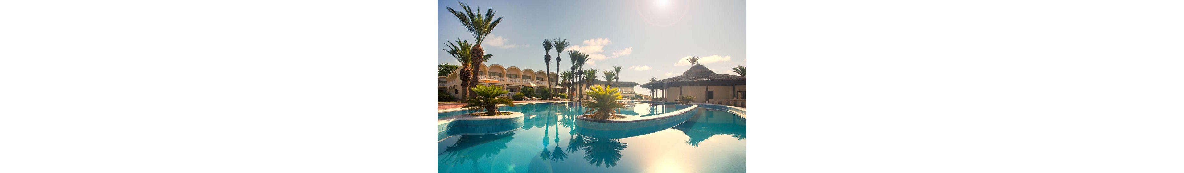 A large, curved swimming pool reflects the bright sun and clear sky at a tropical resort. Palm trees and small landscaped islands decorate the pool, creating a relaxing vacation atmosphere. Resort buildings with balconies line one side, while a thatched-roof bar or pavilion sits on the opposite side. The warm lighting and calm water suggest a peaceful, sunny day ideal for leisure and relaxation.