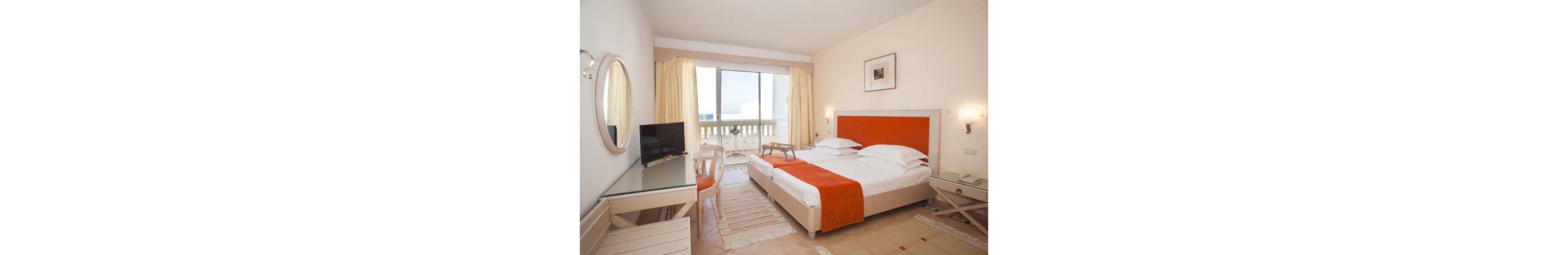 A neatly arranged hotel room features a double bed with white linens and orange accents. The space includes a glass-topped desk with a chair, a wall-mounted television, and light-colored furniture. Large curtains frame a sliding glass door that opens to a balcony with a balustrade and a distant outdoor view. Soft neutral tones and natural light create a calm, inviting atmosphere suitable for relaxation or work.