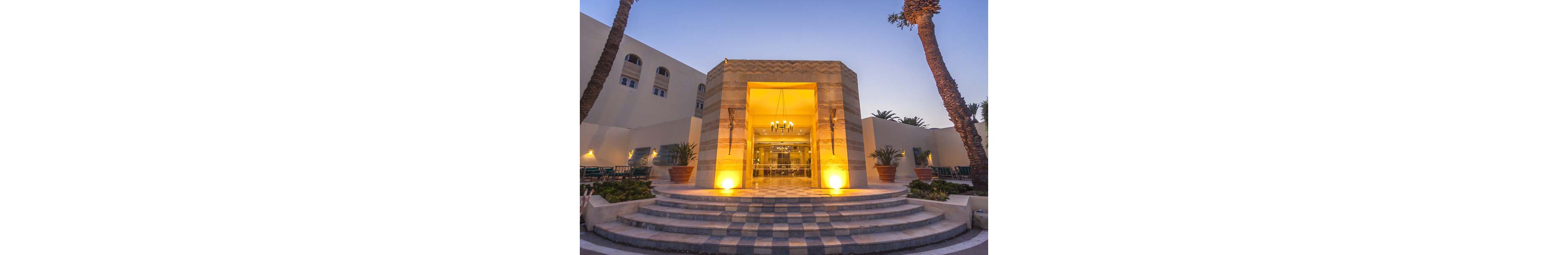 A warmly lit hotel entrance is framed by tall palm trees at dusk. Curved stone steps lead up to a glass doorway, where a chandelier glows inside the lobby. The building features light-colored walls and geometric patterns, creating a modern yet inviting appearance. Soft evening sky tones contrast with the golden lighting, emphasizing the welcoming atmosphere.