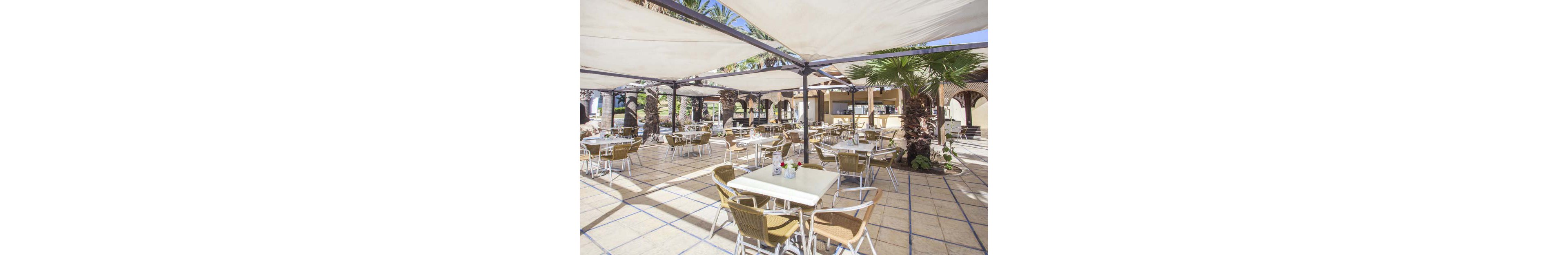 An open-air café terrace features multiple tables and wicker chairs arranged neatly under large white shade canopies. The setting appears bright and sunny, with palm trees integrated into the tiled patio. A bar or service counter is visible in the background, suggesting a resort or hotel environment. The overall scene feels relaxed and inviting, ideal for outdoor dining or refreshments.