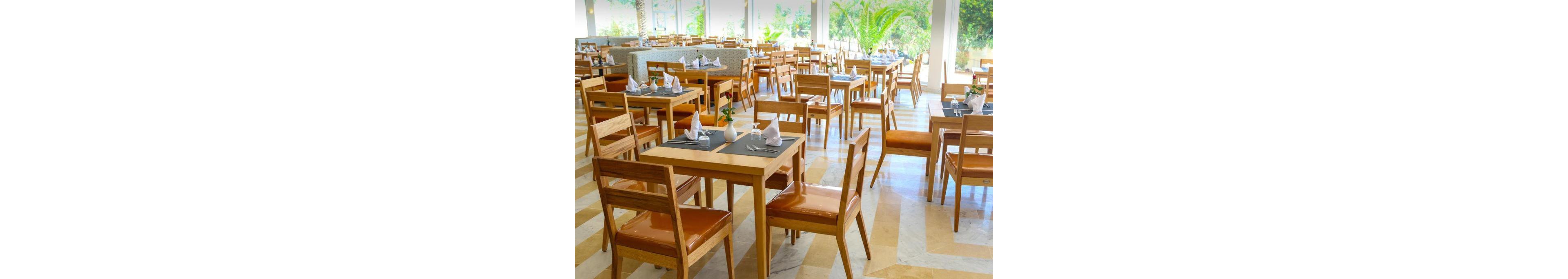 A spacious restaurant dining area features rows of wooden tables and chairs neatly arranged. Each table is set with placemats, folded napkins, and small decorative vases, ready for guests. Large windows and glass doors let in abundant natural light, highlighting the warm tones of the wood and the patterned floor. Green plants and trees are visible outside, adding a fresh, inviting atmosphere to the modern interior.