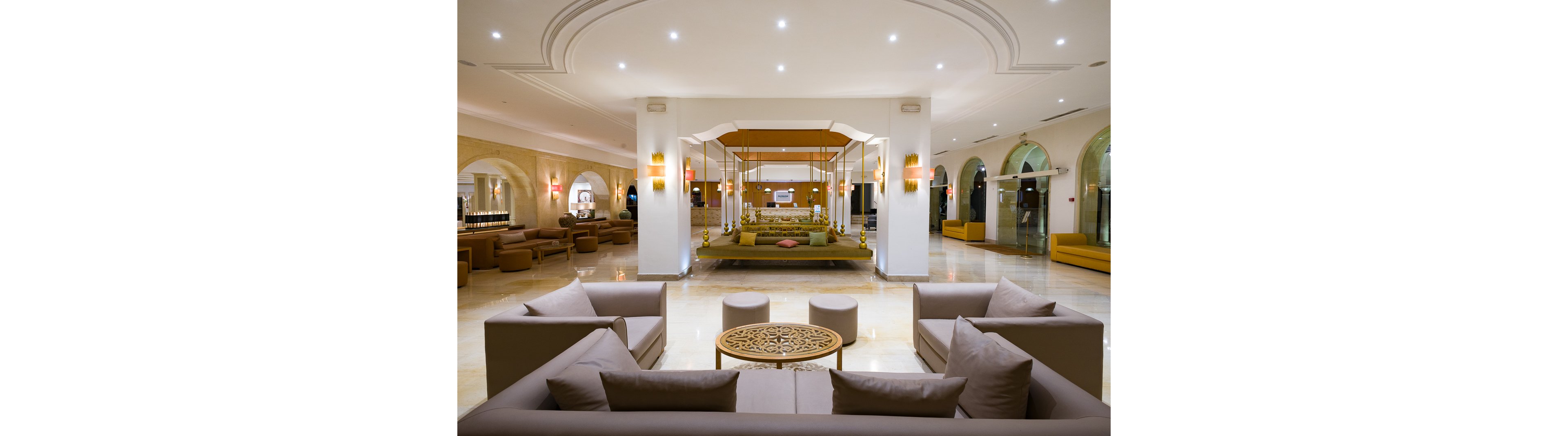 A spacious, modern hotel lobby features neutral-toned sofas arranged around a decorative round table in the foreground. At the center of the space, an elegant swing-style seating area is highlighted by warm lighting and floral accents. The polished floor reflects recessed ceiling lights, creating a bright and inviting atmosphere. Arched windows and additional seating areas line the sides of the lobby, enhancing the sense of openness and comfort.