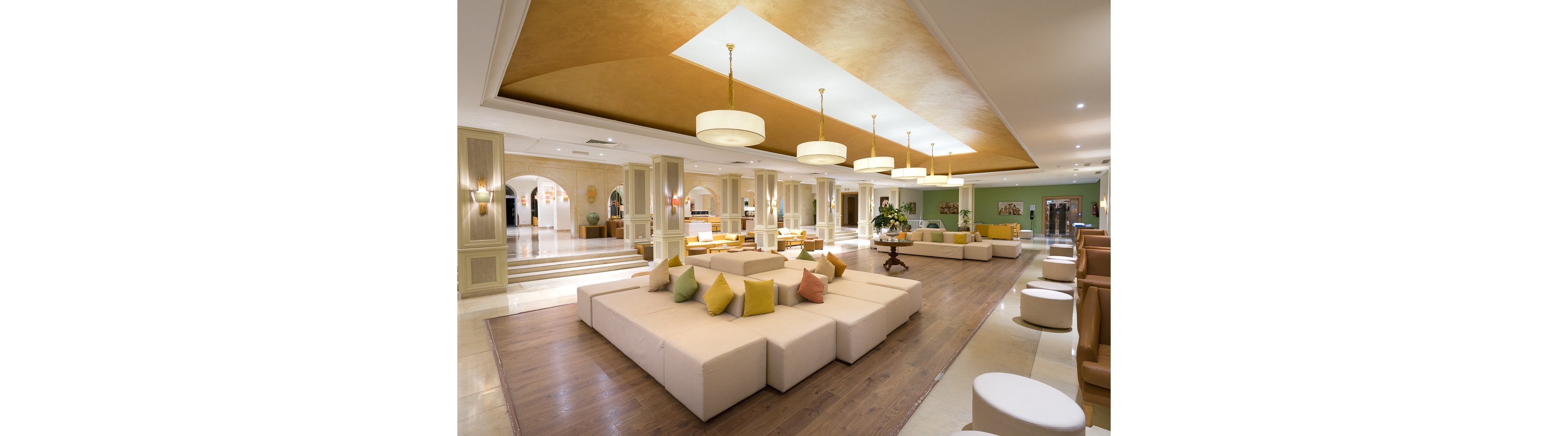 A large, contemporary hotel lobby features modular cream-colored sofas arranged around a central seating area. Soft yellow, green, and orange cushions add color to the neutral furniture and wooden flooring. Circular pendant lights hang from a recessed ceiling, casting warm light across the space. In the background, arches, steps, and additional lounge areas create depth and a welcoming atmosphere.