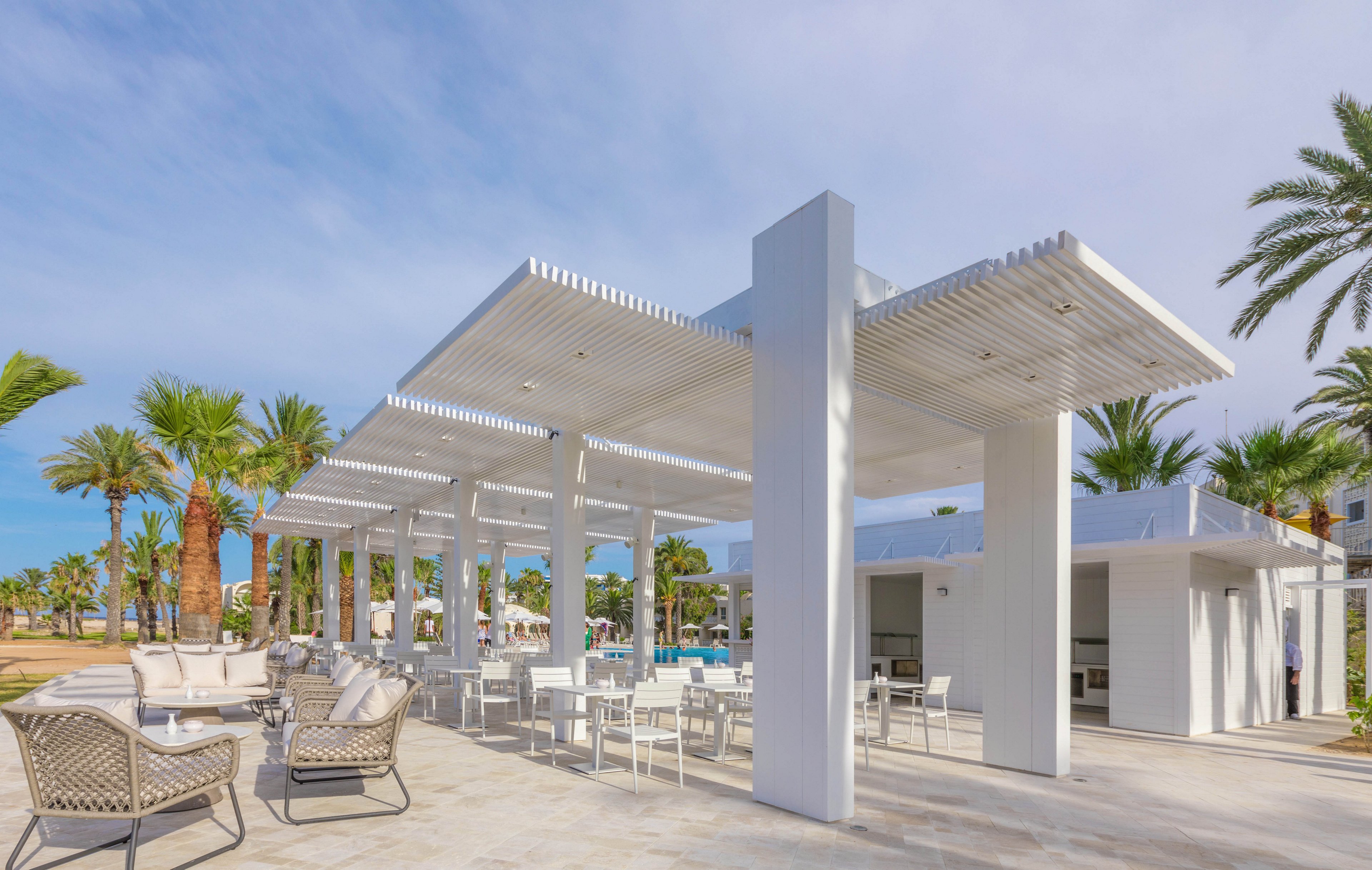 An open-air bar with a sleek white pergola structure stands in a sunny resort setting. White tables, chairs, and lounge seating are arranged on a spacious patio under the shade. Tall palm trees and manicured landscaping surround the area, suggesting a tropical or desert oasis. The bright daylight and clear sky create a relaxed, vacation atmosphere.
