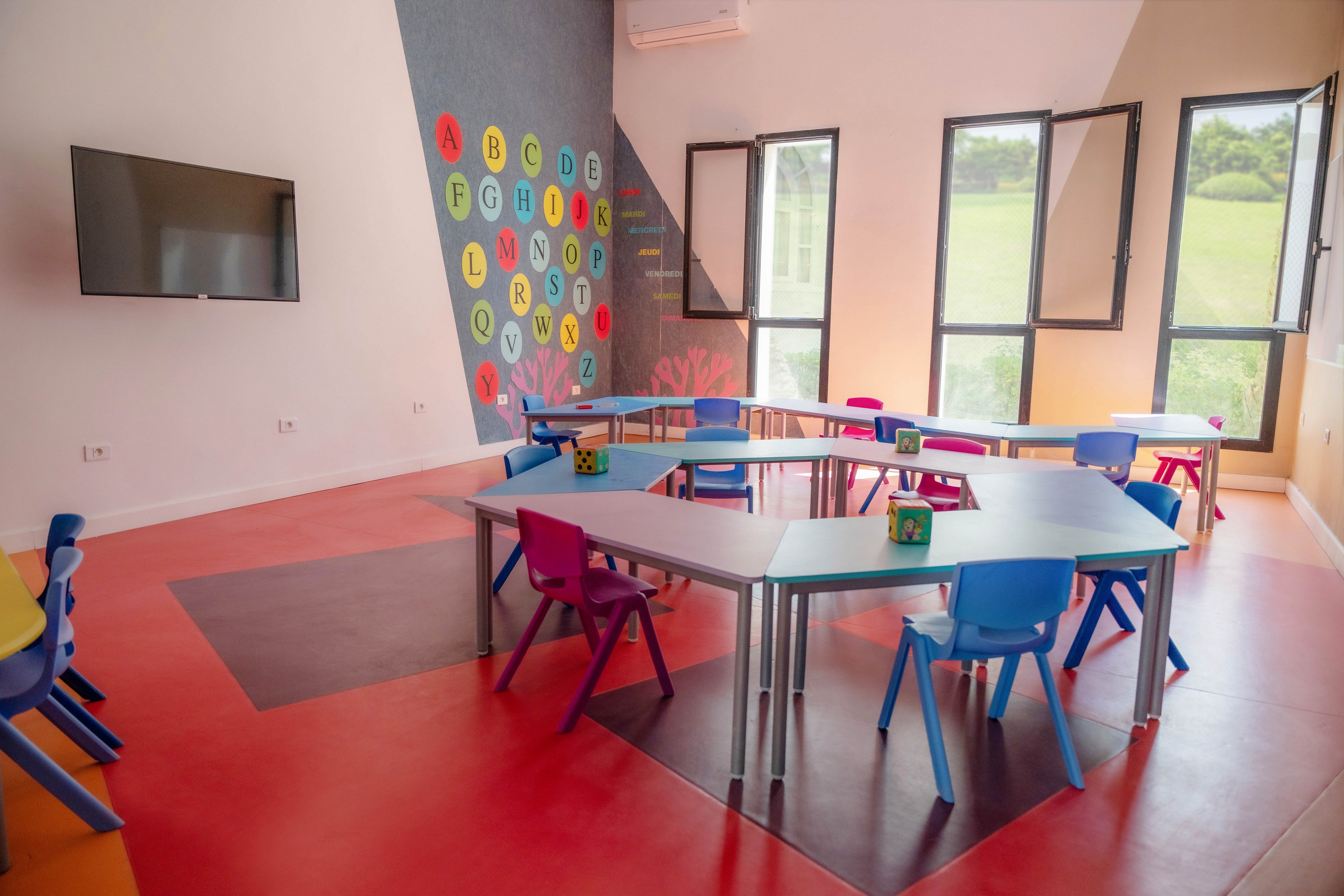 A bright preschool classroom features colorful hexagonal tables and small plastic chairs arranged in a collaborative layout. The room has large windows letting in natural light and a wall-mounted flat screen on one side. A decorative wall displays the alphabet letters A through Z in vibrant circles, adding a playful learning element. The red and gray flooring and pastel walls create a cheerful, modern educational environment.