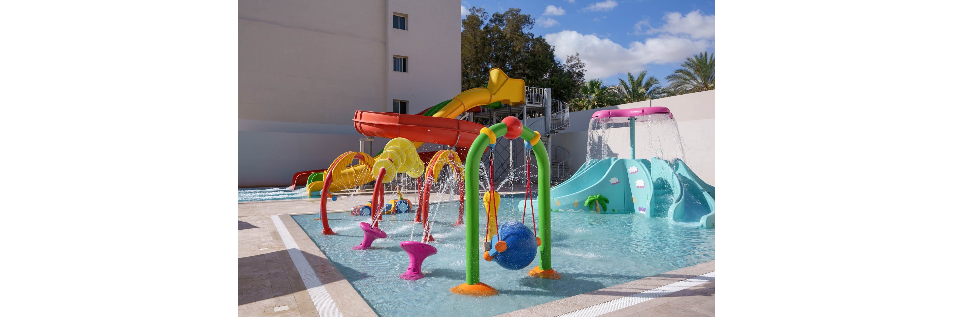 A vibrant children’s splash park features multiple colorful water play structures and slides. Curved spray arches, spinning fountains, and playful shapes spray water into a shallow pool. In the background, a large orange and yellow waterslide curves down from an elevated platform. Palm trees and a light-colored building frame the sunny outdoor setting. The scene suggests a fun, family-friendly recreational area at a resort or water park.
