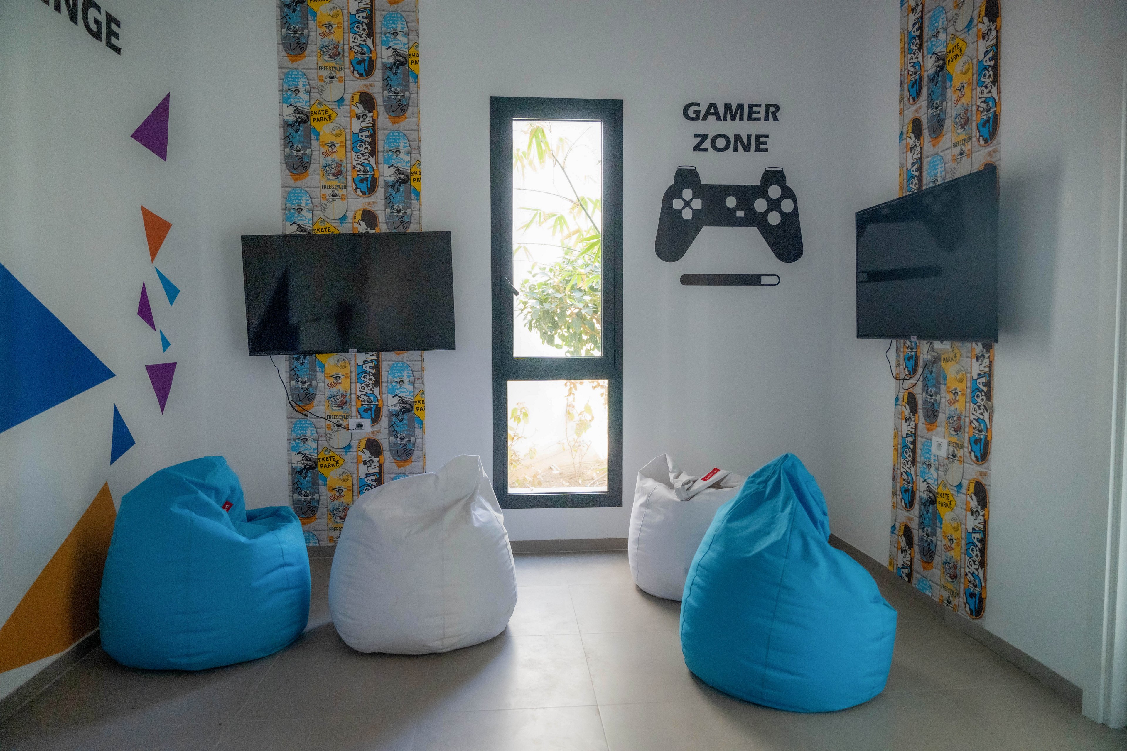 A small gamer zone features multiple bean bag chairs arranged in front of wall-mounted screens. The room has bright blue and white bean bags placed on a light-colored floor. Decorative wall panels with graphic patterns and geometric shapes add a playful touch. A window in the center lets in natural light, and a large controller graphic with the words 'GAMER ZONE' emphasizes the gaming theme.