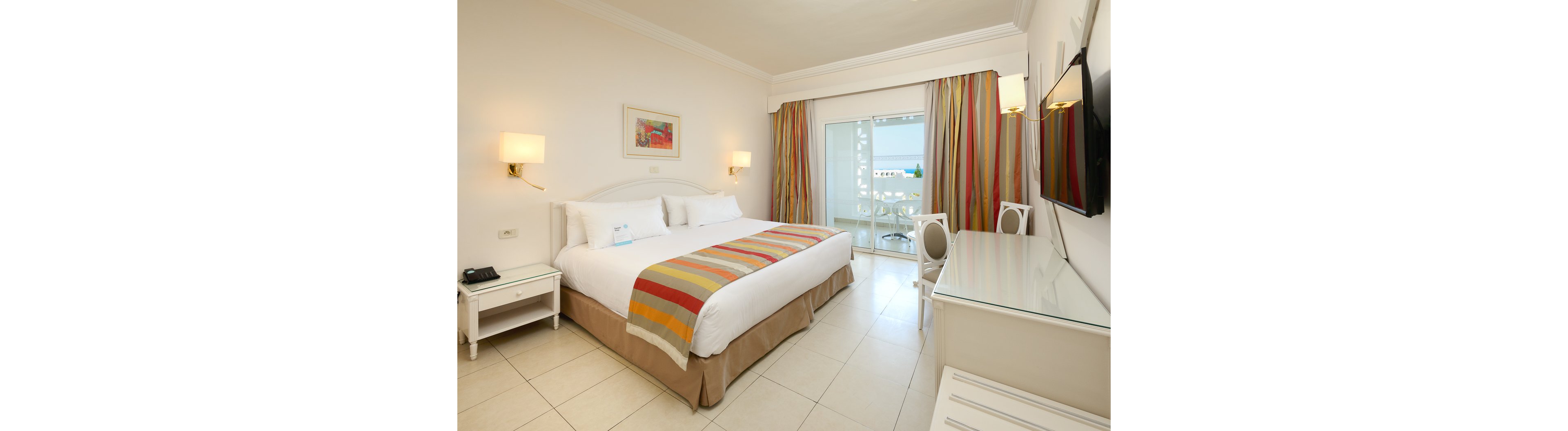 A spacious hotel bedroom features a large bed with white linens and a colorful striped runner. The room includes a bedside table with a phone, a wall-mounted television, and a glass-topped desk with a chair. Floor-to-ceiling curtains in multicolored stripes frame a sliding glass door leading to a balcony. Through the balcony, an outdoor view with blue sky and distant buildings is visible, adding natural light to the clean, modern interior.
