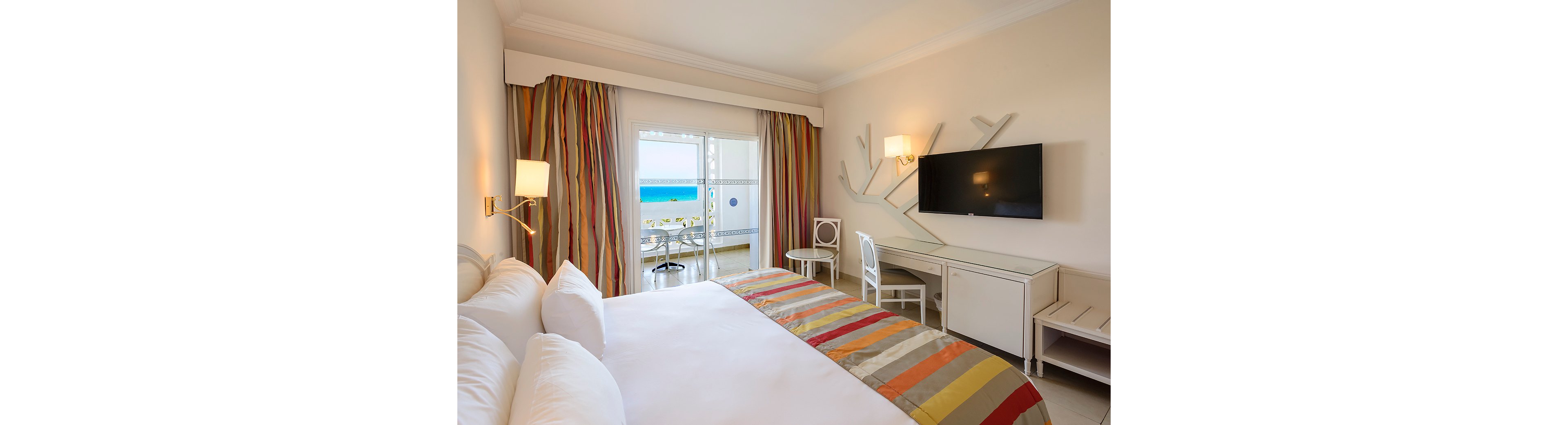 A modern hotel room features a large neatly made bed with colorful striped bedding. The space includes a wall-mounted flat-screen television, white desk, and chairs arranged near a balcony door. Floor-to-ceiling striped curtains frame the glass doors that open to a balcony with a table and chairs overlooking a bright blue ocean. Warm wall sconces and neutral walls create a clean, inviting atmosphere ideal for a beach vacation stay.