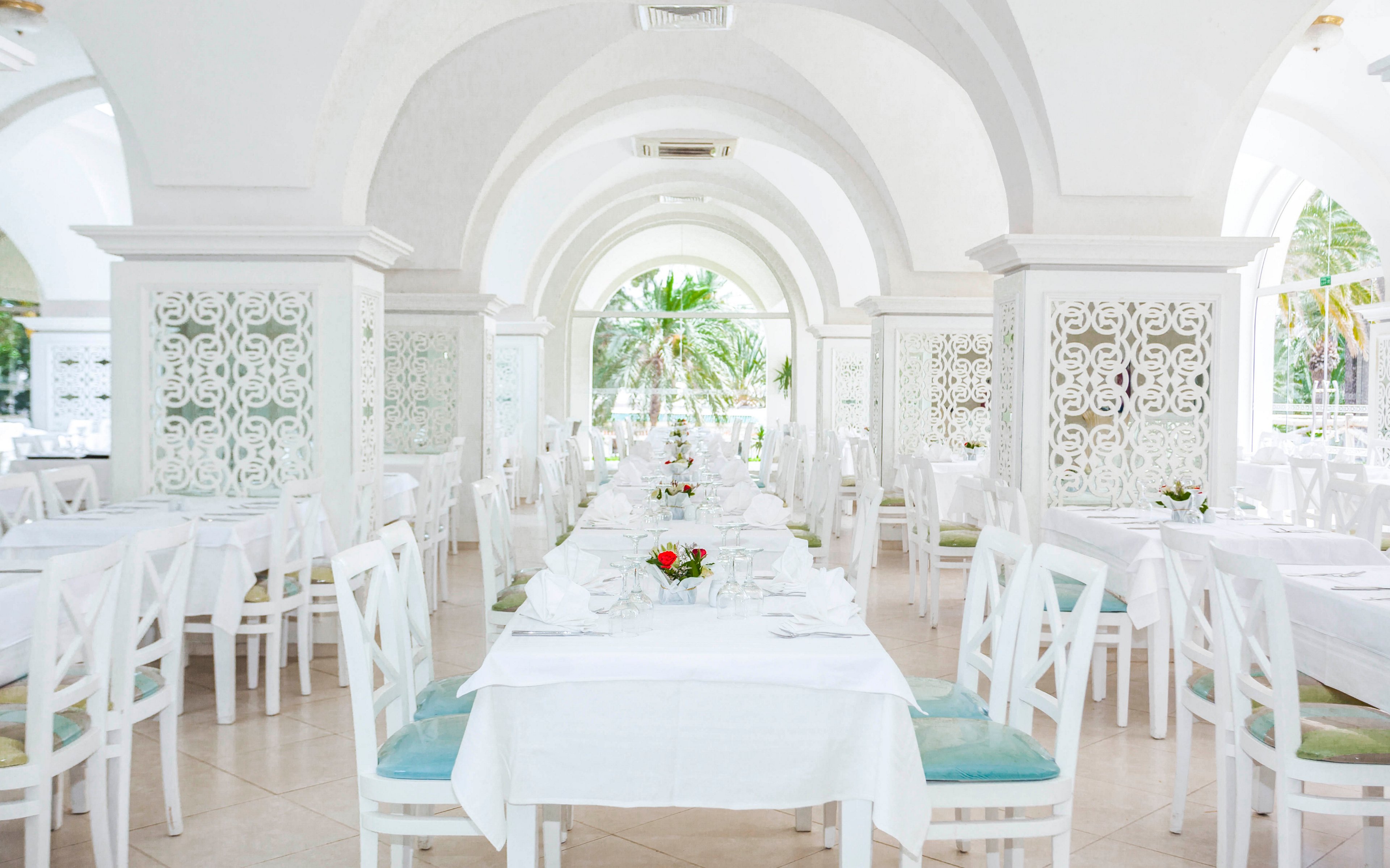 A bright, elegant restaurant interior features long banquet tables arranged in rows. White tablecloths, neatly folded napkins, and small floral centerpieces create a refined dining setting. Arched ceilings and decorative lattice panels add architectural interest to the space. Large openings at the far end reveal greenery and palm trees outside, enhancing the airy, tropical atmosphere.