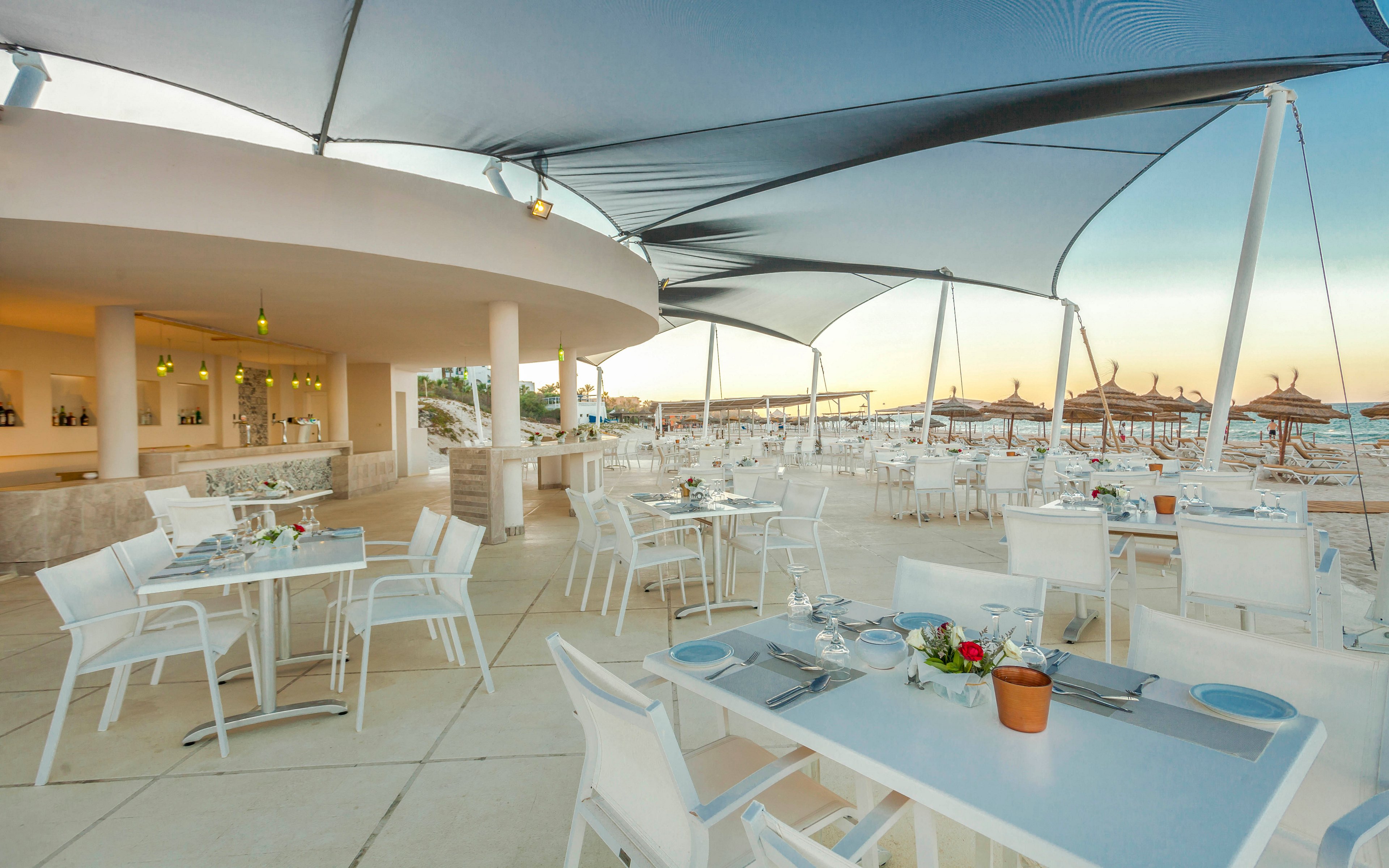 An open-air restaurant terrace is set beside a sandy beach, featuring rows of white tables and chairs under modern sail-like shade canopies. The bar area in the background is softly lit, with bottles and glassware neatly arranged. Beach umbrellas and lounge chairs line the shoreline, creating a relaxed seaside atmosphere at sunset. The scene feels calm and inviting, ideal for outdoor dining by the ocean.