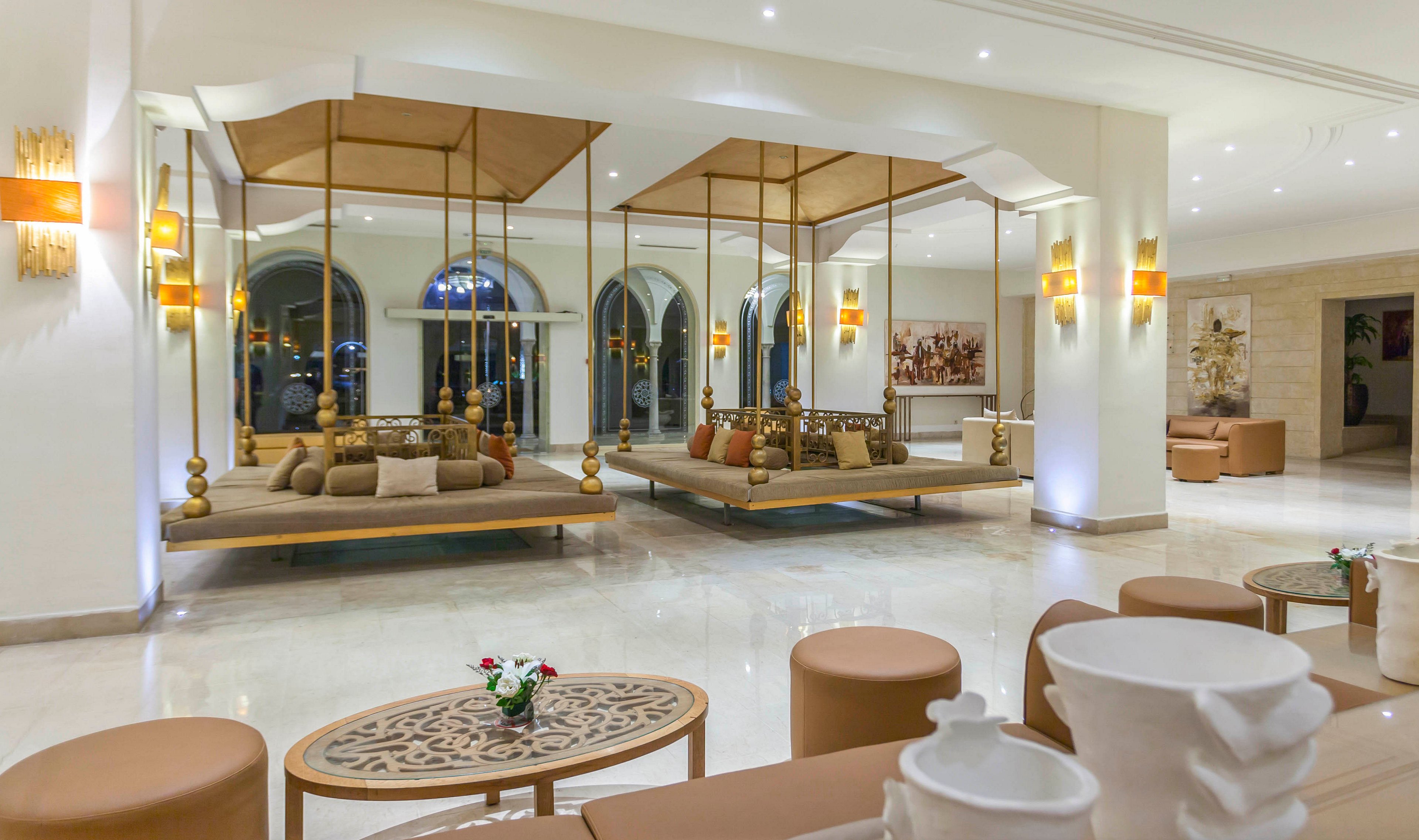 A spacious, contemporary hotel lobby features large suspended lounge beds with plush cushions and pillows. The seating area is framed by tall metal supports and positioned in front of arched windows that reflect the interior lighting. Warm wall sconces and recessed ceiling lights create a soft, inviting glow across the polished floor. In the foreground, low tables with floral arrangements and sculptural white decor add to the sophisticated, modern ambiance.