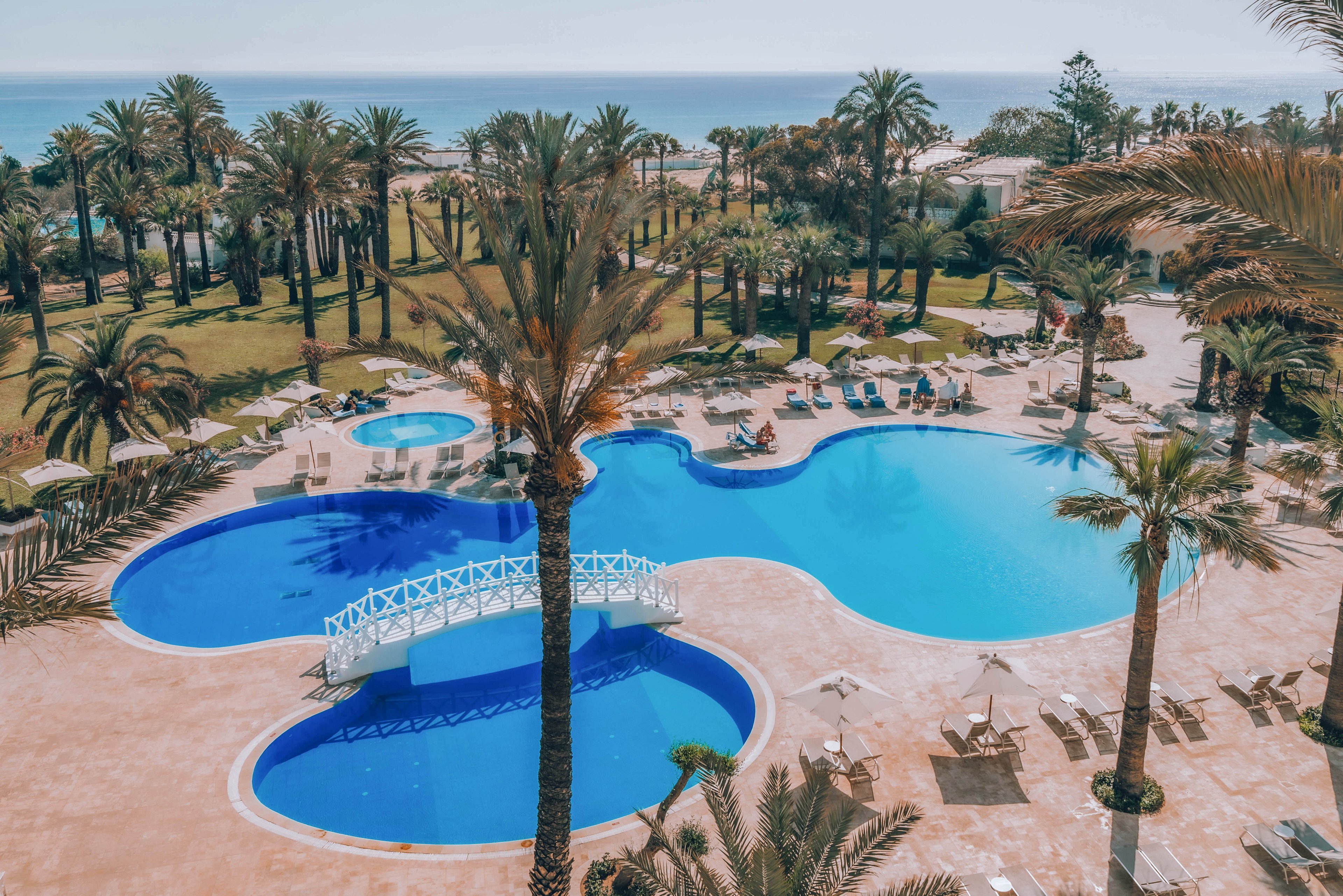 An expansive resort swimming pool is surrounded by tall palm trees and sun loungers. A small white footbridge crosses one section of the pool, adding a decorative focal point. The scene includes multiple pool areas with curved shapes and a tiled deck with umbrellas and chairs. In the background, a lush garden and the distant sea create a tranquil vacation atmosphere under clear skies.
