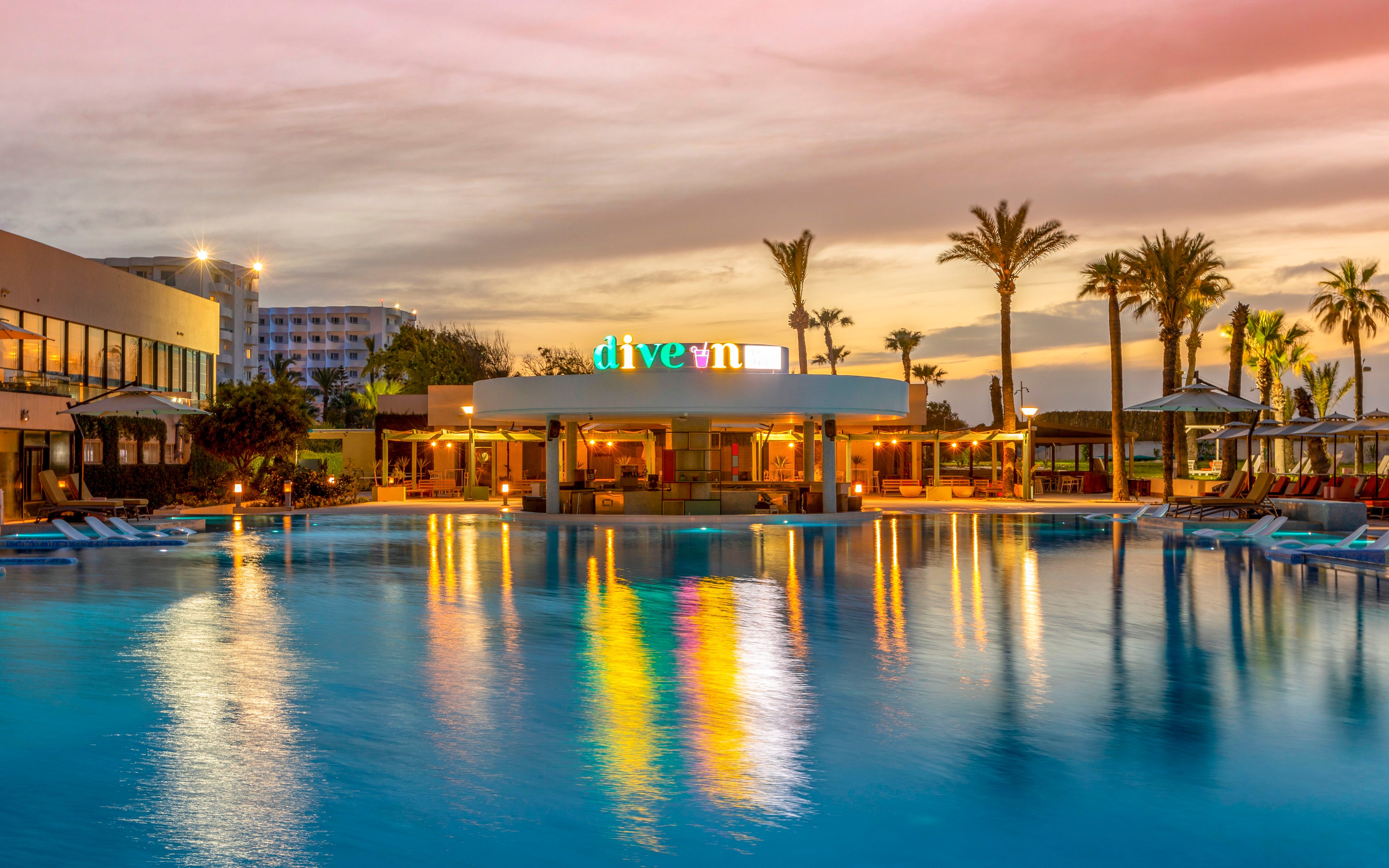 A large resort swimming pool is shown at sunset, reflecting colorful lights from a central bar area with the text "dive inn" glowing above it. Palm trees and lounge chairs surround the pool, creating a relaxed vacation atmosphere. The sky is painted in warm hues of orange, pink, and purple, enhancing the tranquil mood. Soft lighting from the buildings and cabanas adds to the inviting, tropical setting.