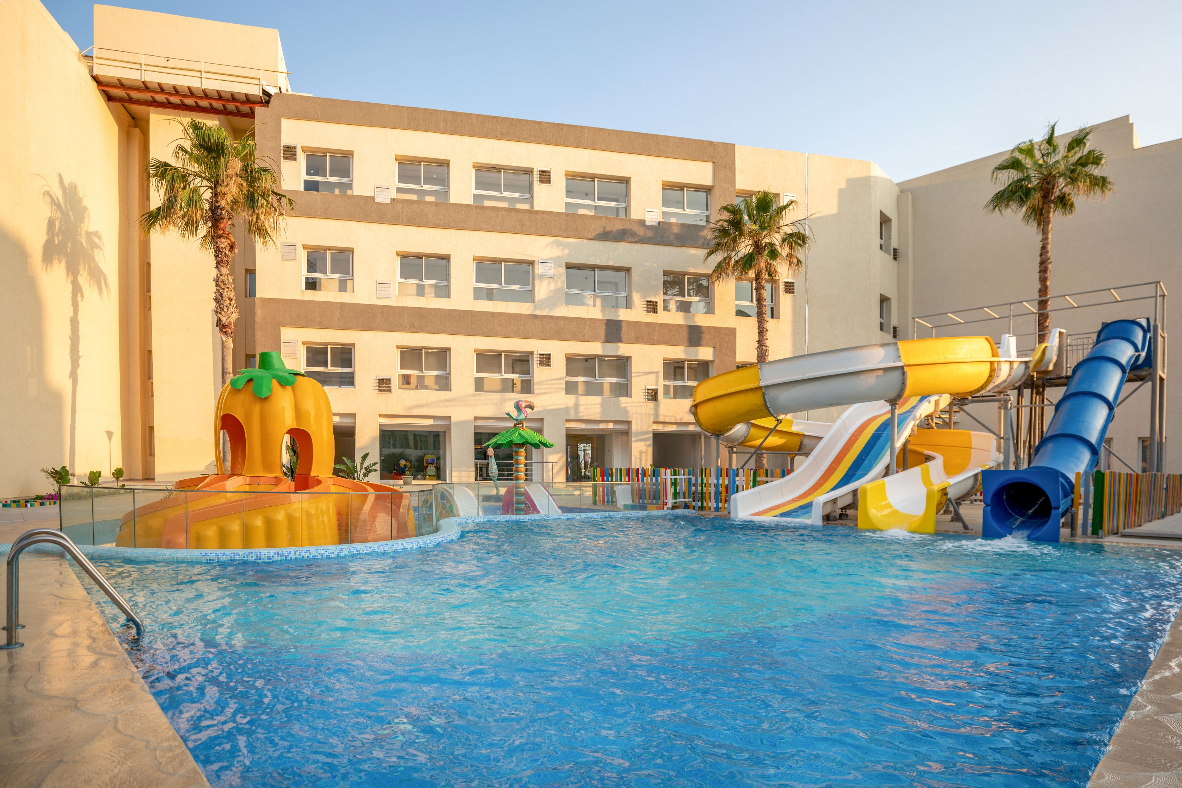 An outdoor swimming pool with colorful waterslides is set in front of a multi-story hotel or apartment building. The pool area features playful structures, including a pumpkin-shaped slide and a large yellow and blue tube slide. Palm trees frame the scene, and the warm sunlight suggests a late afternoon or early evening setting. The clear blue water and bright colors create a fun, family-friendly atmosphere ideal for vacation or leisure.