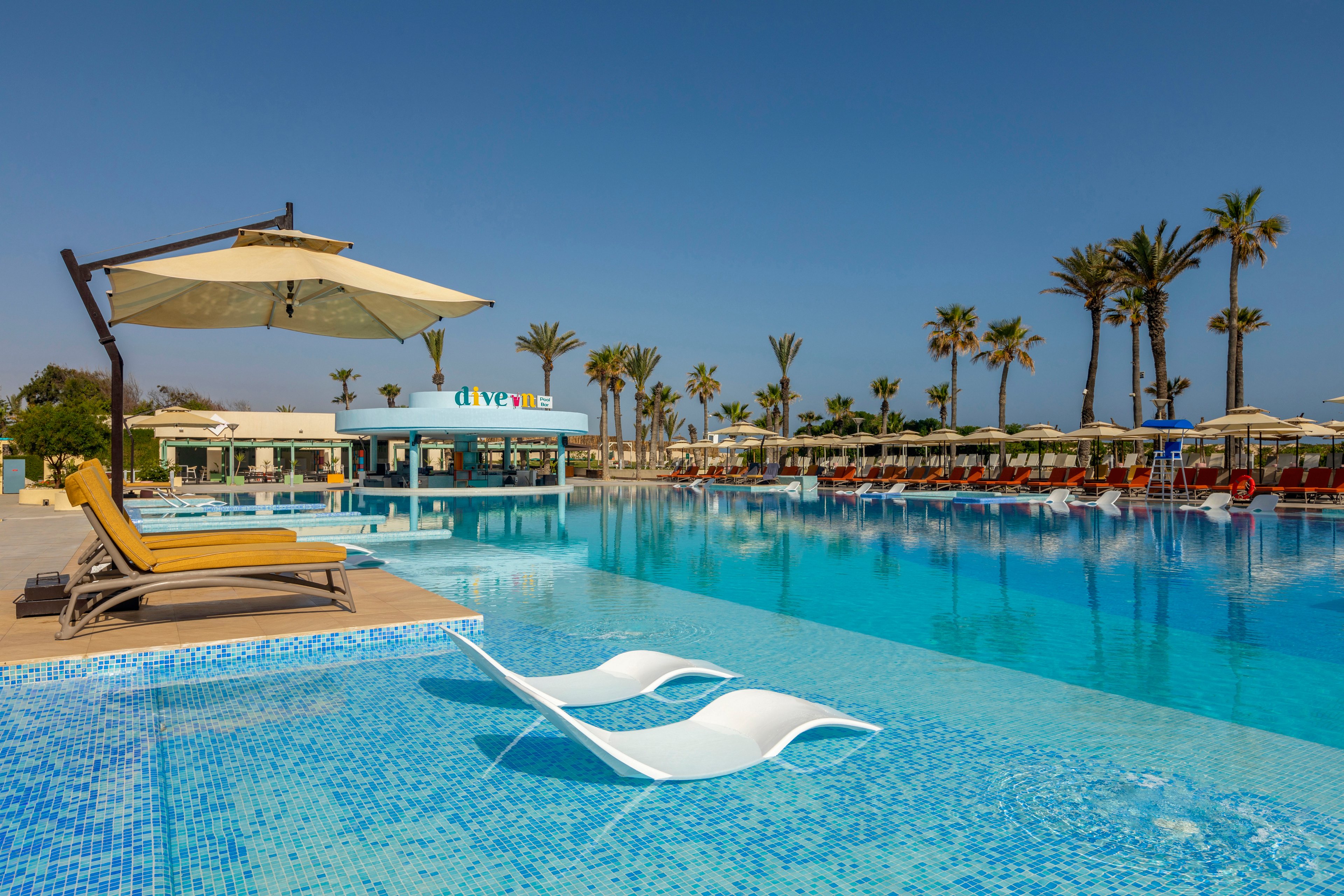 A large resort swimming pool stretches out under a clear blue sky, featuring shallow in-water loungers in the foreground. Yellow cushioned sunbeds sit beneath a wide umbrella on a wooden deck beside the pool. In the background, rows of sun loungers and umbrellas line the pool edge, with tall palm trees adding a tropical feel. A poolside bar building with the visible text "dive in" stands at the far side of the water. The scene conveys a calm, sunny vacation atmosphere with bright blue and sandy tones.