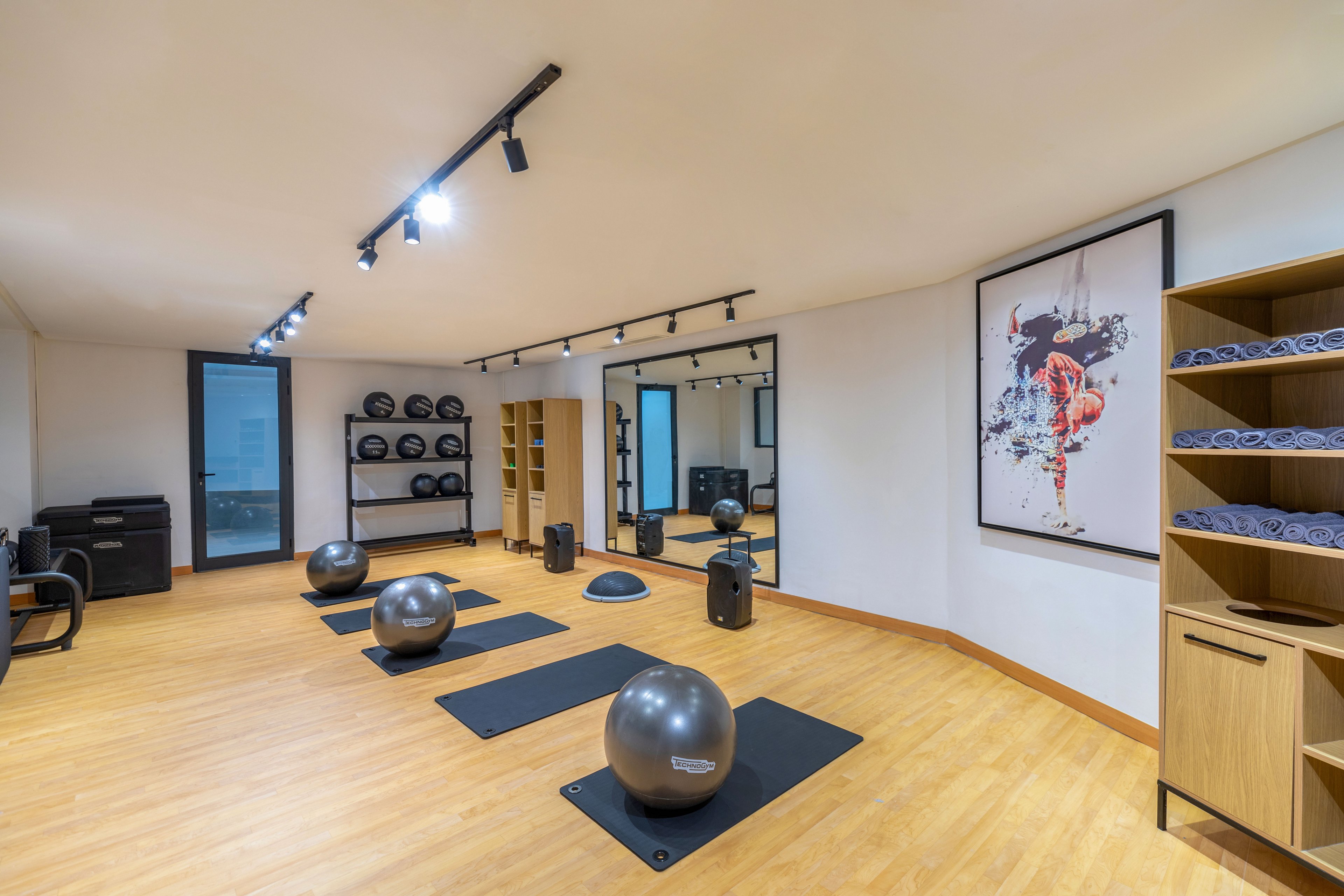 A bright, modern fitness studio features several exercise balls and mats neatly arranged on a wooden floor. The room includes shelving units with rolled towels and stacked weights, suggesting a space for yoga, pilates, or functional training. Track lighting illuminates the area, and a large wall mirror expands the sense of space. A colorful abstract artwork adds visual interest to the otherwise minimalist interior.