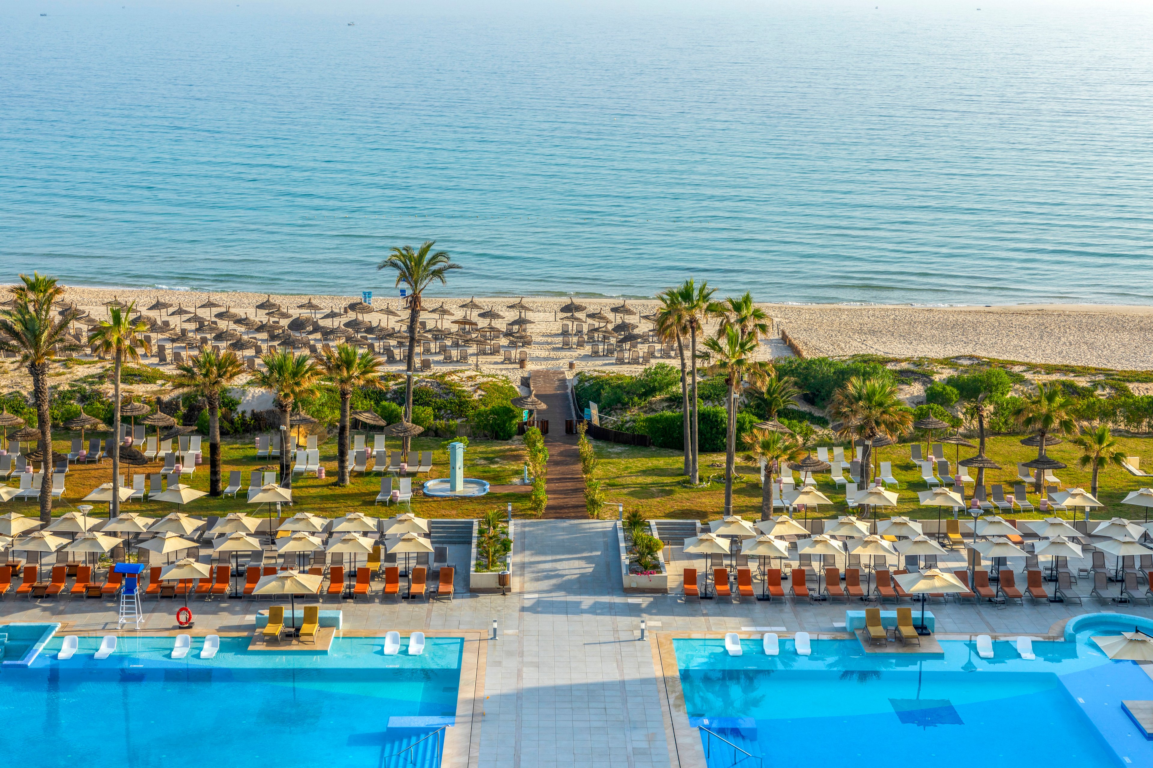A large seaside resort features two bright blue swimming pools in the foreground overlooking a sandy beach. Neatly arranged sun loungers and umbrellas line the pool deck and extend down toward the shoreline. Palm trees and manicured lawns create a tropical atmosphere between the resort and the sea. Calm ocean water stretches to the horizon under clear skies, suggesting a relaxing vacation setting.