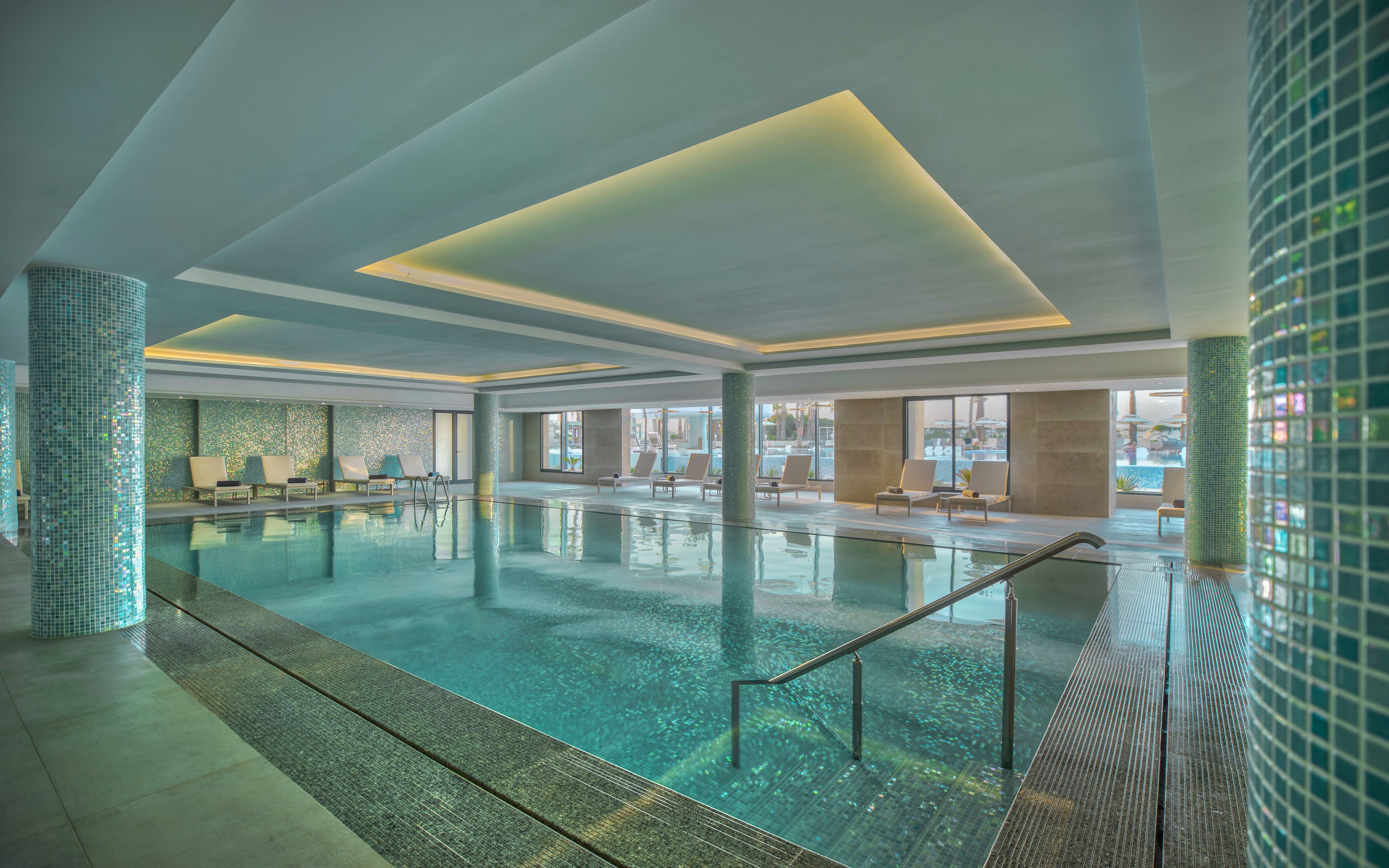 A spacious indoor swimming pool is featured in a contemporary hotel or spa setting. The pool is surrounded by tiled columns and walls, with a metal handrail leading into the water. Lounge chairs line one side of the pool near large windows that let in natural light. Soft ceiling lighting and blue-green mosaic tiles create a calm, luxurious atmosphere.
