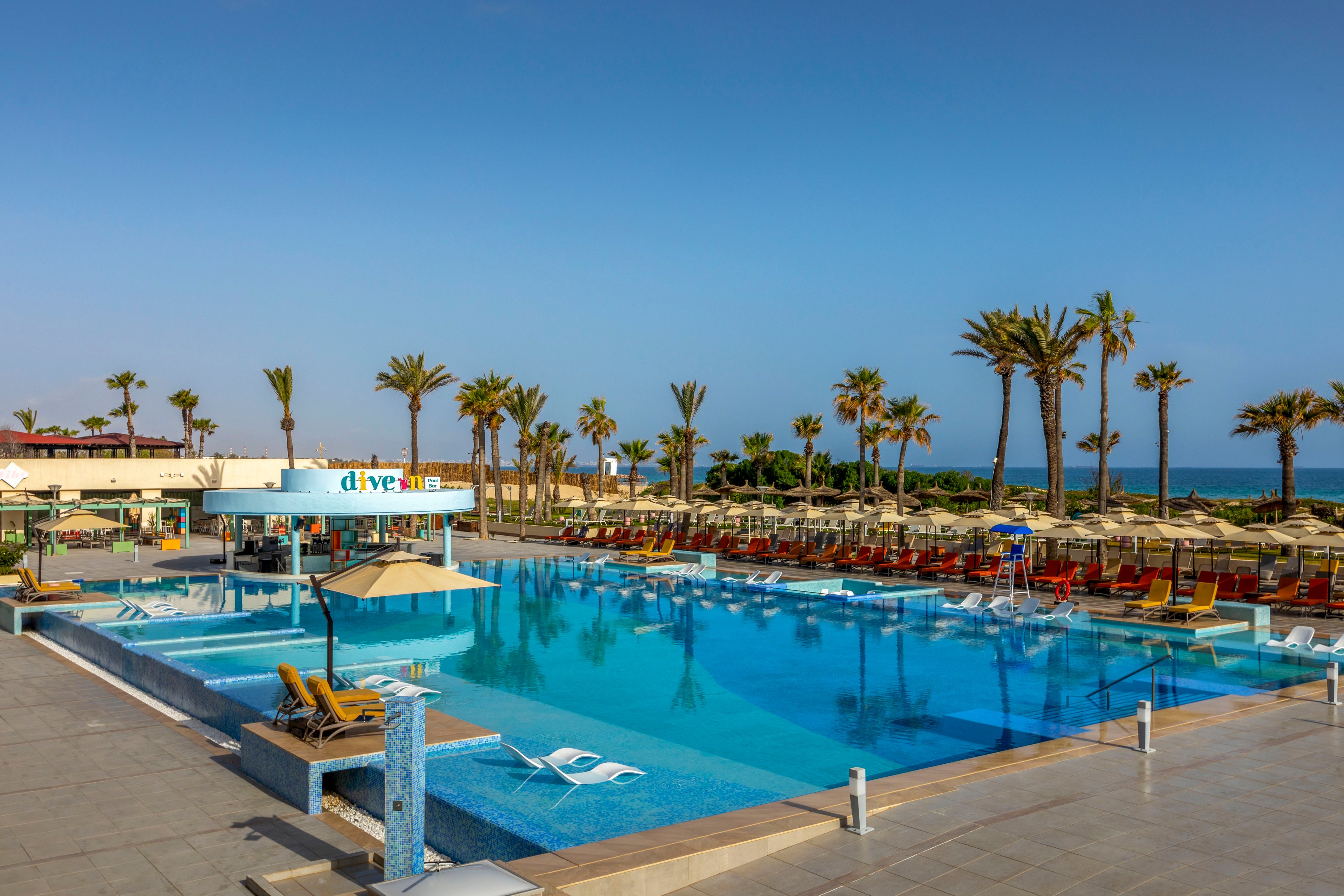 A large resort-style infinity pool stretches out toward the ocean under a clear blue sky. Sun loungers and umbrellas line the pool deck, with palm trees framing the scene. A swim-up bar structure sits within the pool, and shallow lounging areas with submerged chairs are visible in the foreground. The calm water reflects the surrounding palm trees and bright, sunny weather, creating a relaxing vacation atmosphere.
