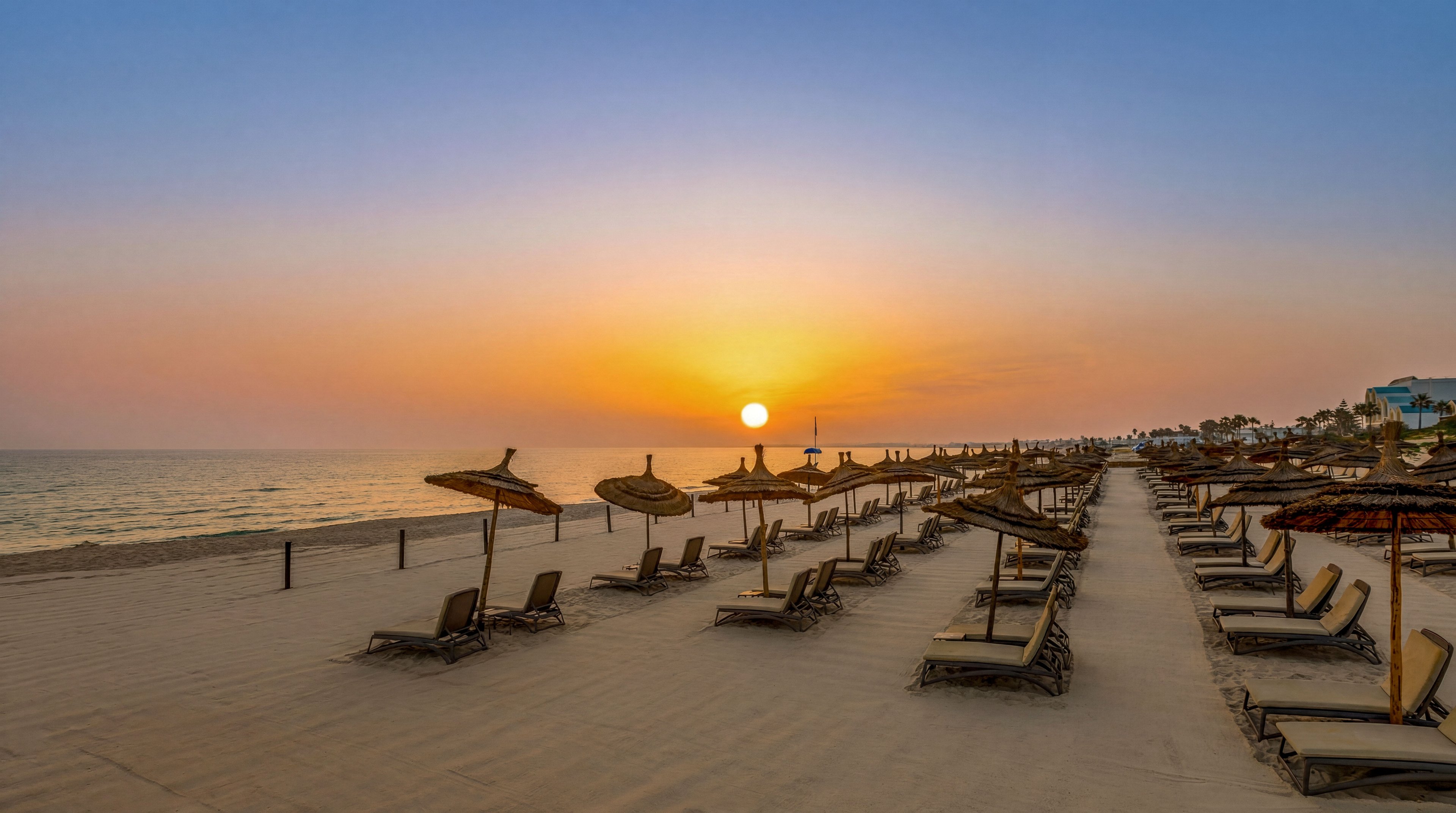 A tranquil beach scene shows rows of empty lounge chairs facing the ocean at sunset. Straw parasols line the sandy shore, creating a symmetrical pattern toward the horizon. The sun is low in the sky, casting warm golden light across the calm sea and beach. The atmosphere feels peaceful and serene, suggesting a quiet moment at a seaside resort.
