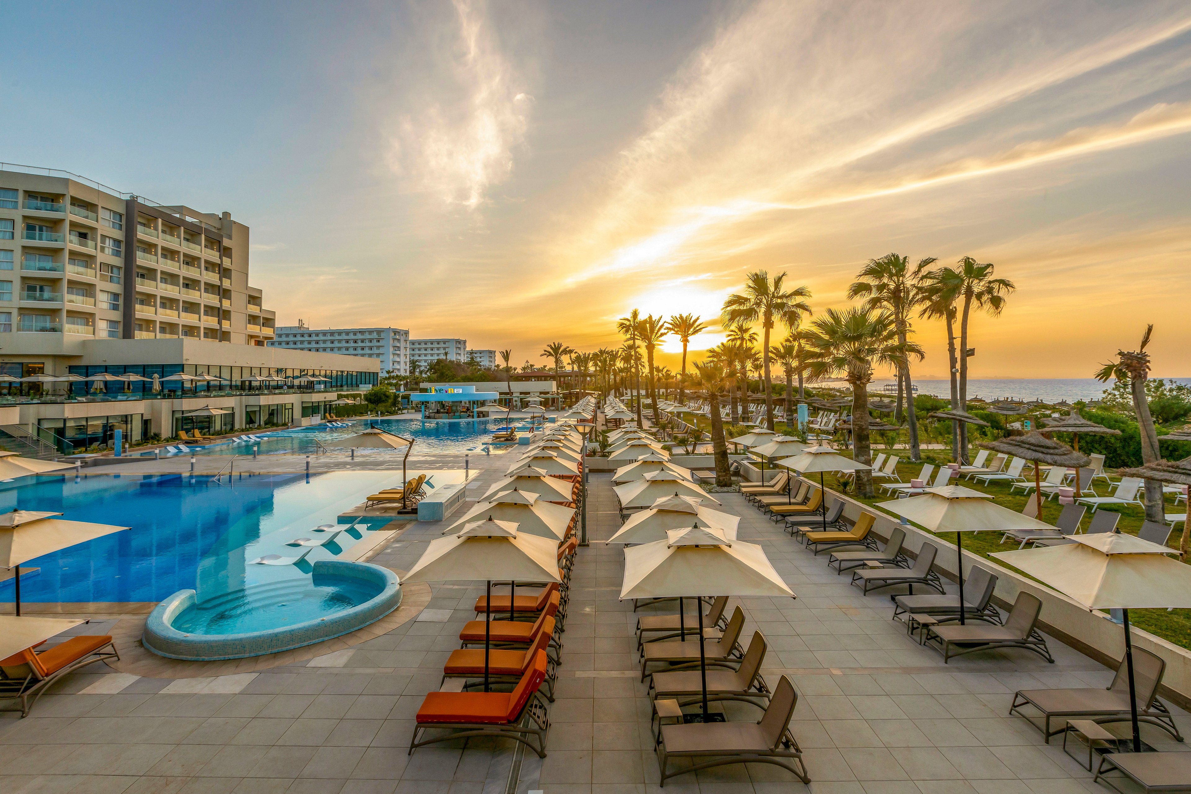 A large resort swimming pool is lined with rows of sun loungers and umbrellas, facing the ocean at sunset. Modern multi-story hotel buildings rise on the left, while palm trees frame the view toward the beach and horizon. The sky glows with warm golden light and soft clouds, reflecting on the calm blue water. The scene evokes a relaxing vacation atmosphere with ample seating and spacious poolside terraces.