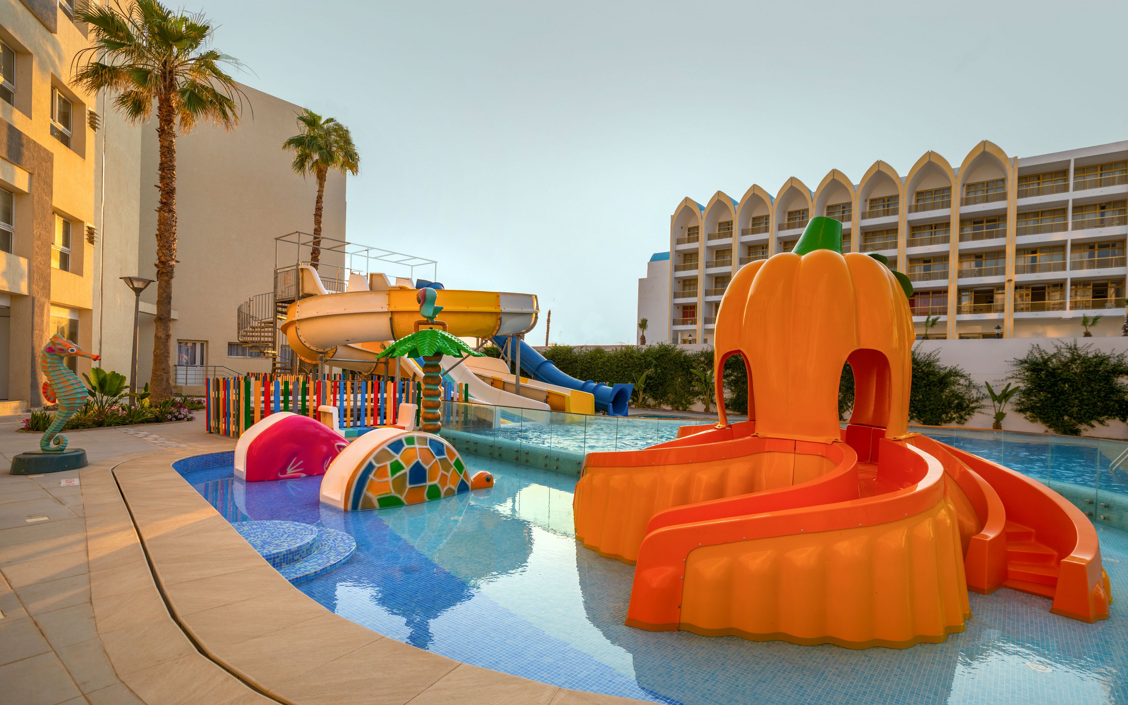 A vibrant children’s water playground features bright orange, yellow, and multicolored slides shaped like playful characters and objects. The structures sit in a shallow pool beside a modern hotel building with balconies and palm trees. The scene is captured at sunset or early evening, giving the sky a warm, soft glow. The overall atmosphere is fun, family-friendly, and inviting for kids on vacation.