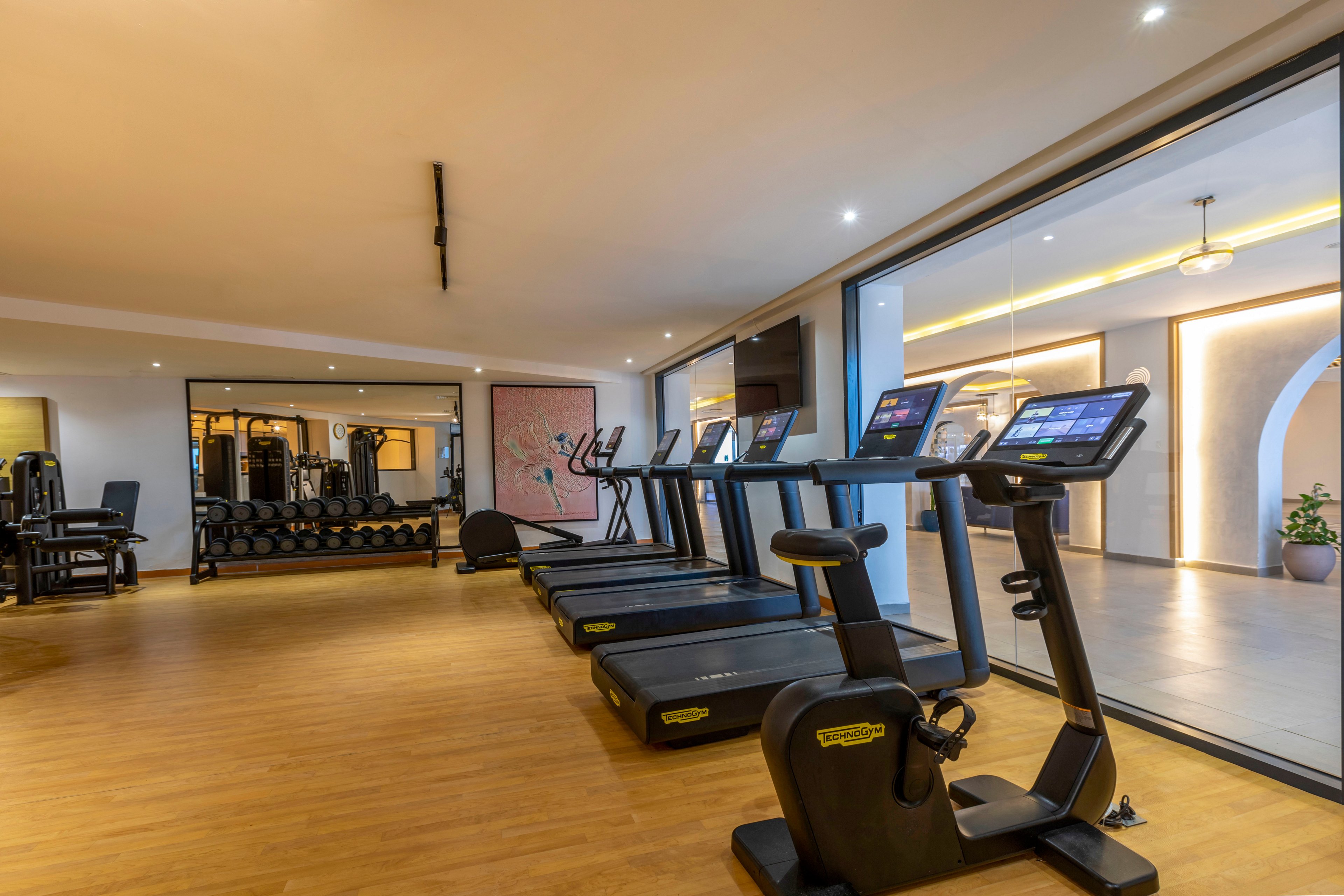 A spacious modern gym features a row of treadmills aligned on a wooden floor. Large mirrors on the far wall reflect additional fitness equipment and create a sense of depth. Floor-to-ceiling glass separates the cardio area from a bright hallway with warm lighting and arches. The overall scene is clean, organized, and ready for indoor workouts.