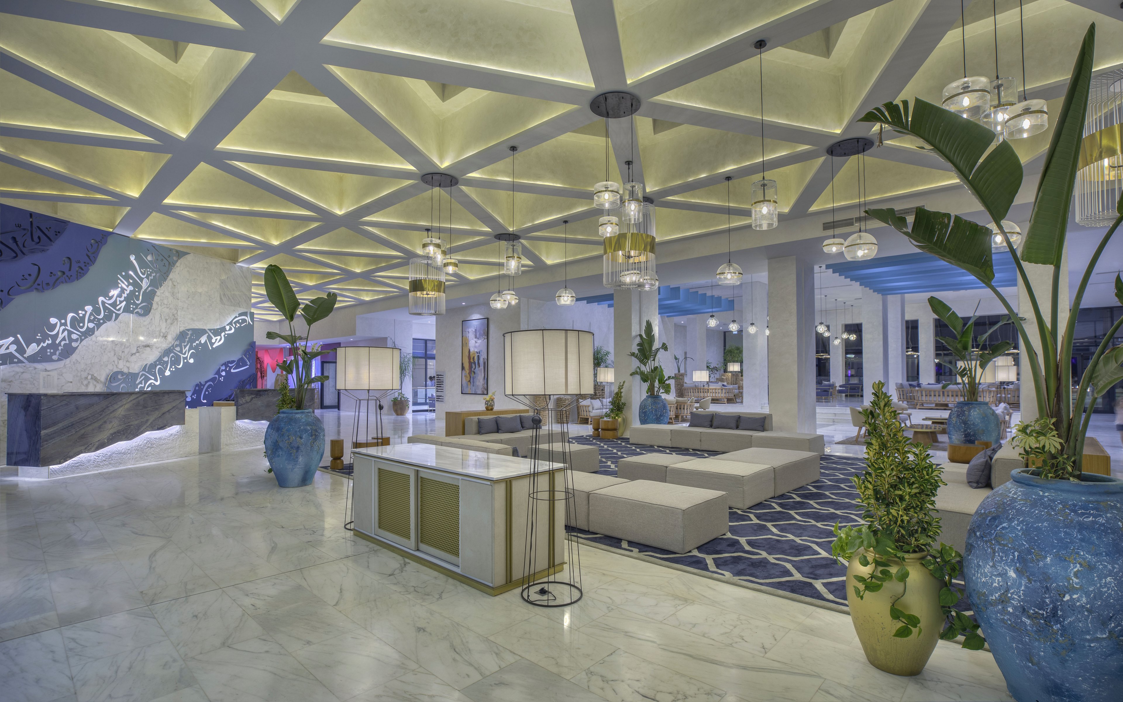 Spacious contemporary hotel lobby featuring elegant lounge seating and decorative lighting. The interior showcases a geometric coffered ceiling with warm illumination and large pendant lamps. Blue and white tones dominate the space, complemented by patterned rugs and marble flooring. Oversized ceramic planters with lush green plants add a touch of nature to the sophisticated setting.