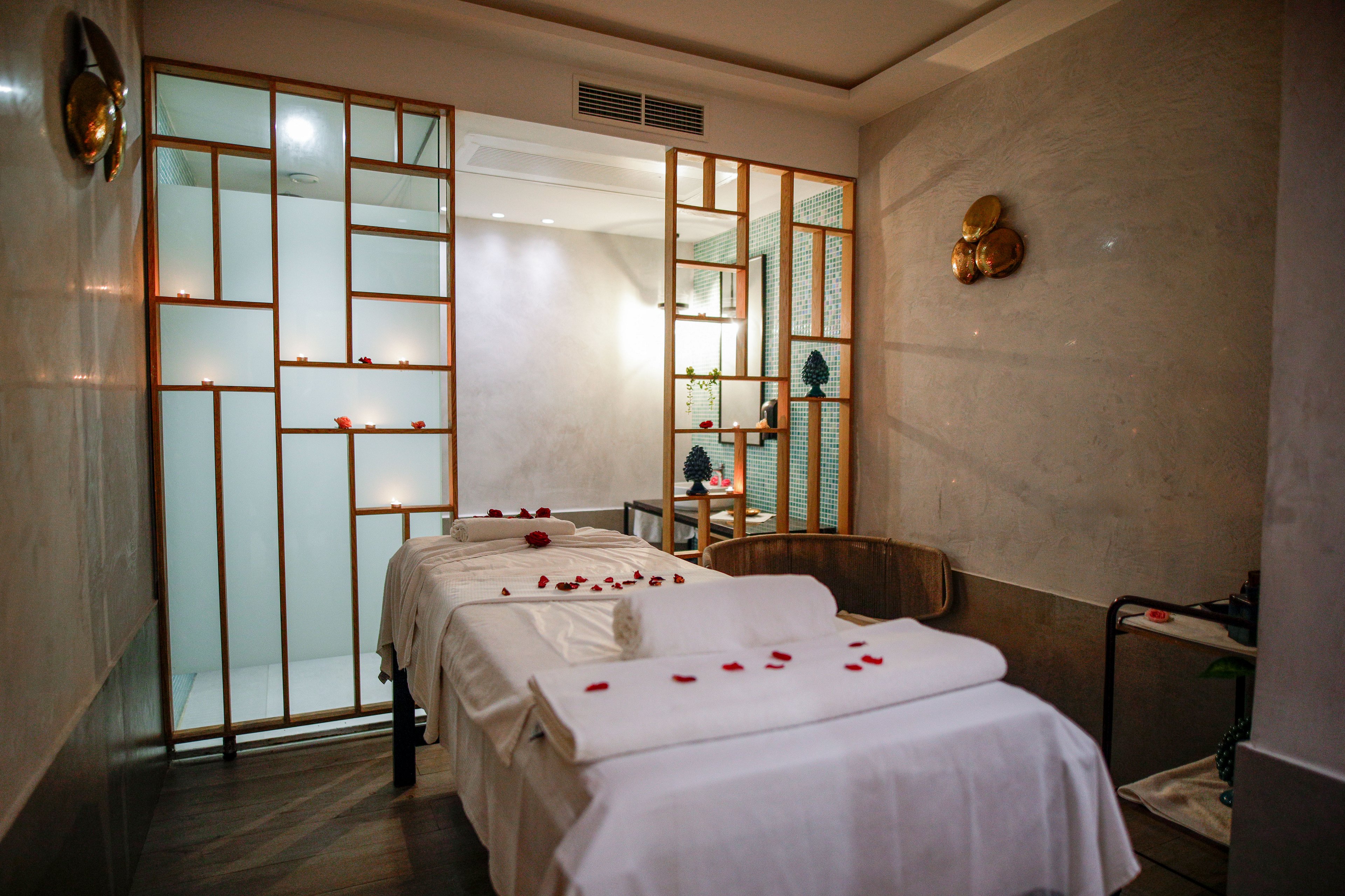 A serene spa treatment room features two massage tables covered in crisp white linens. Rolled towels and scattered red rose petals create a romantic, relaxing atmosphere. Warm lighting reflects off the light-colored walls and decorative metal room divider. Minimalist decor and soft textures emphasize comfort and tranquility in this wellness setting.