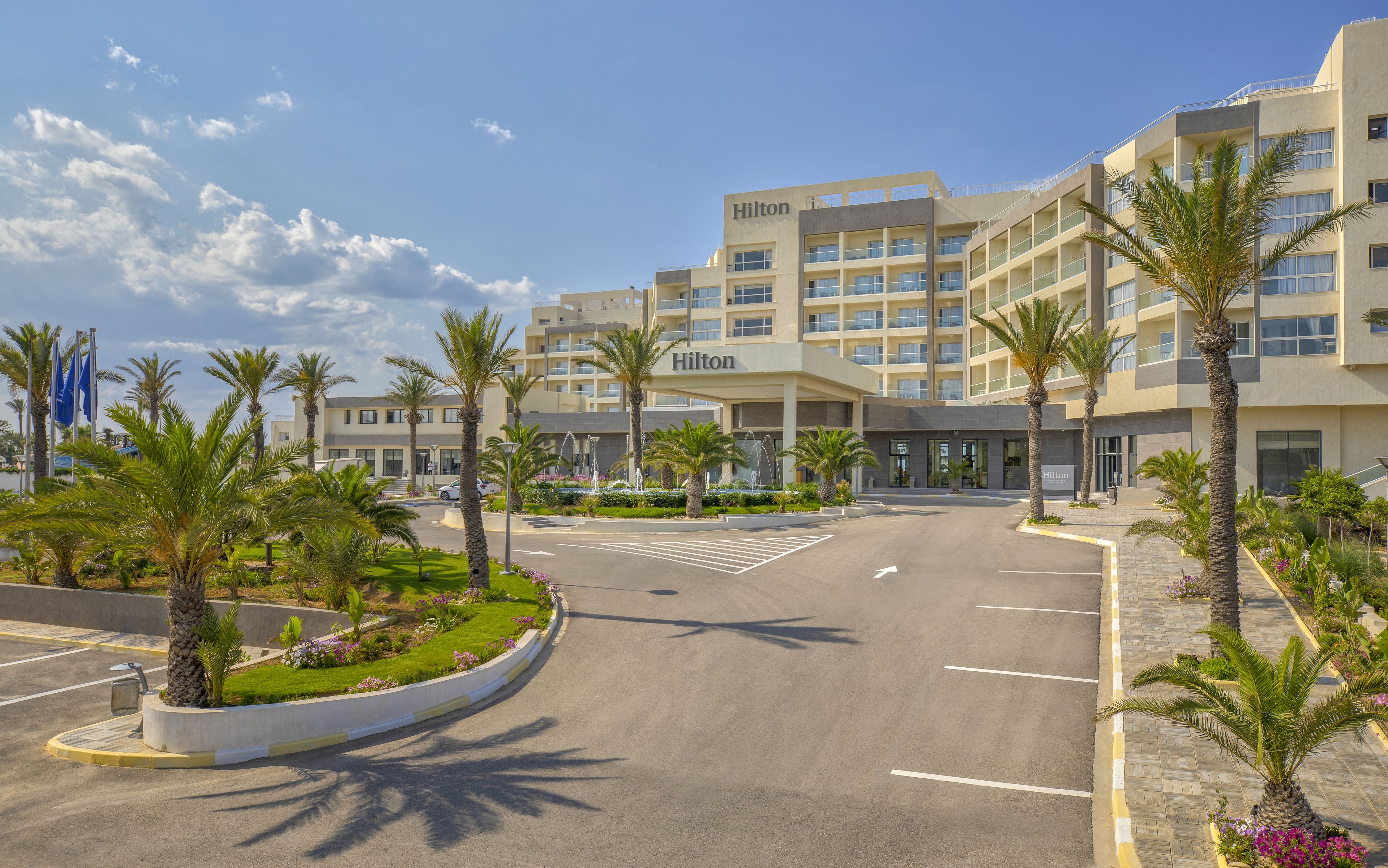 A large modern hotel complex stands prominently with multiple stories and balconies. The entrance is approached by a wide driveway lined with palm trees and landscaped greenery. Clear skies with scattered clouds create a bright, inviting atmosphere. The scene suggests a coastal or resort destination focused on leisure and relaxation.