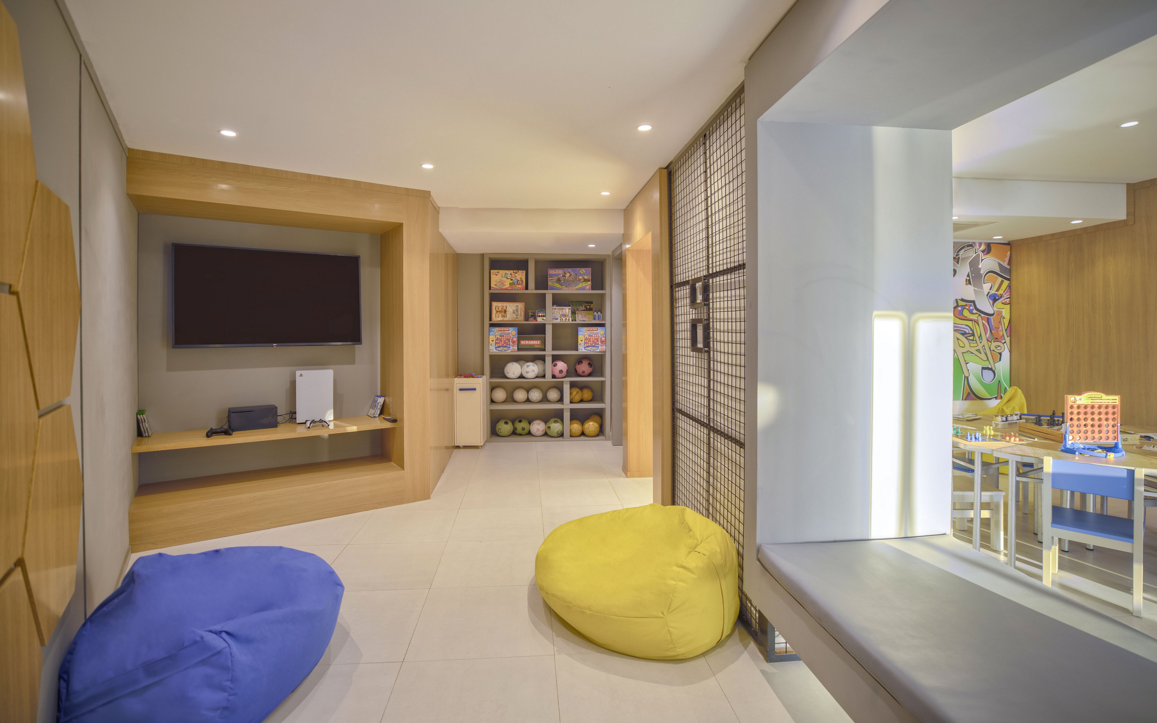 A contemporary playroom features a wall-mounted television and media console on one side. Shelves in the background are filled with toys, books, and sports balls, creating a family-friendly atmosphere. Bright bean bag chairs and a cushioned bench provide casual seating. A connected area with a table and chairs suggests a space for kids’ games and creative activities, all lit by warm recessed lighting.