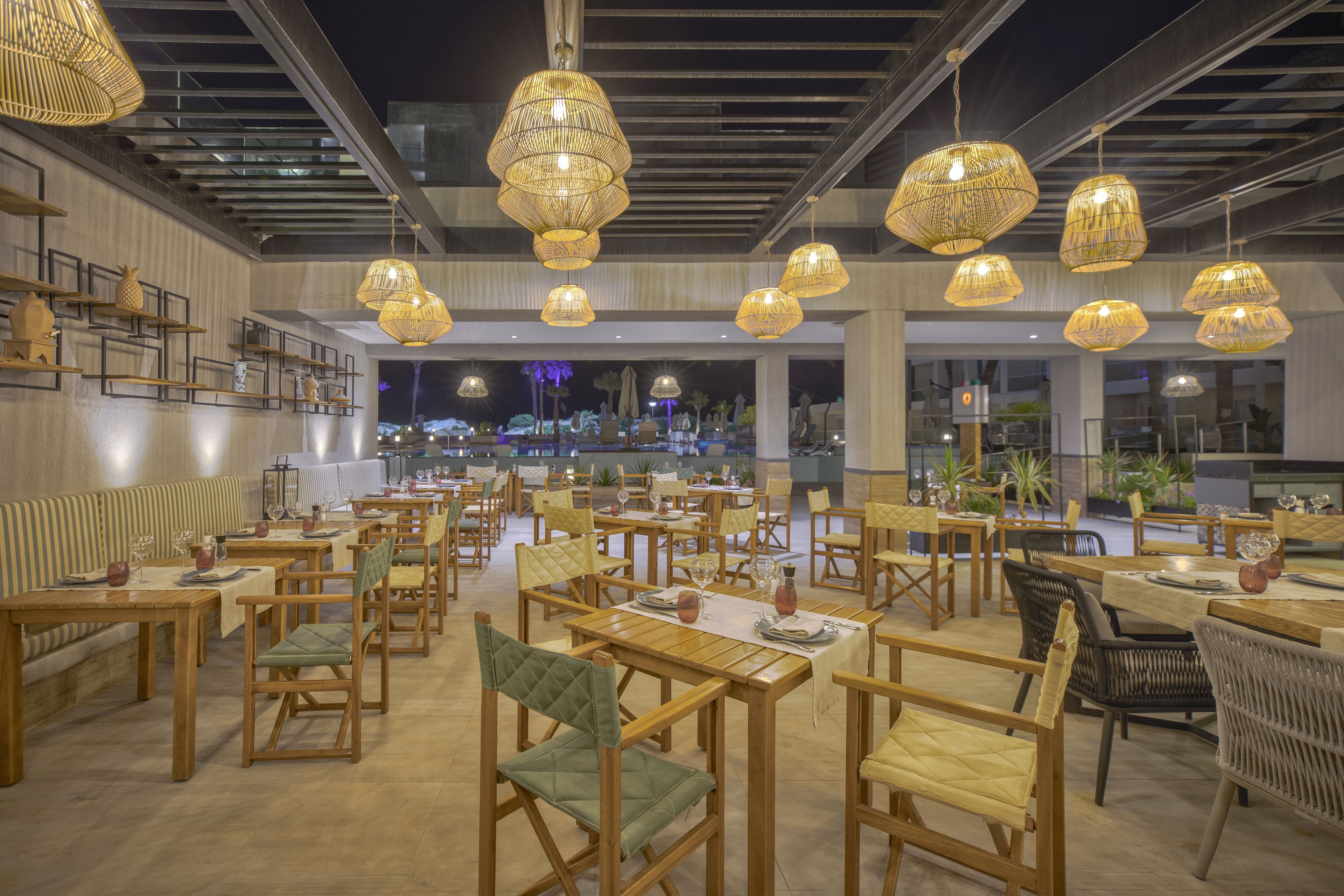 Contemporary restaurant interior featuring wooden tables and cushioned chairs arranged in neat rows. The open-air space overlooks a cityscape at night through large windows. Woven pendant lamps hang from the ceiling, casting warm ambient light across the dining area. Neutral tones, natural materials, and potted plants create a relaxed, inviting atmosphere.