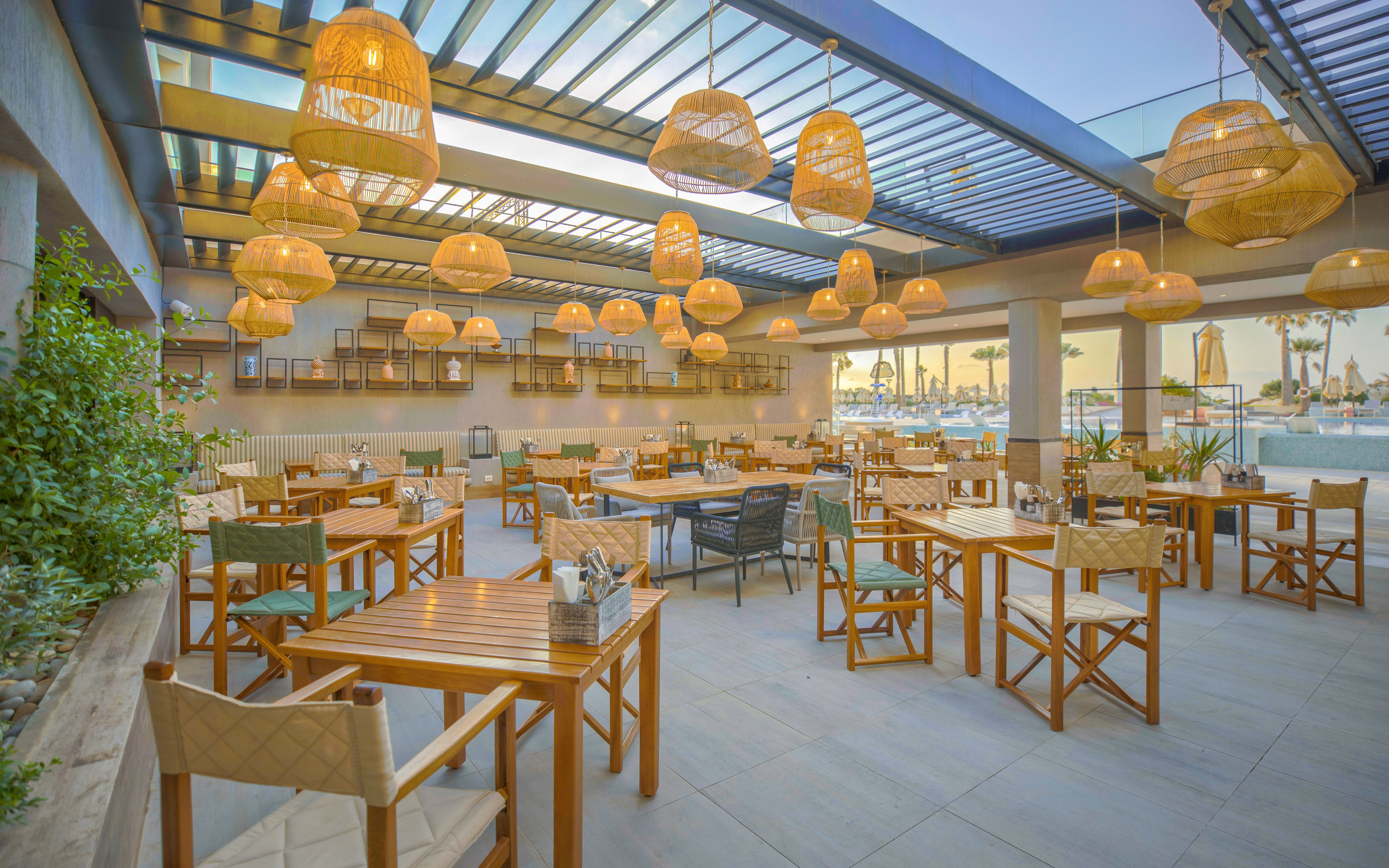 Spacious outdoor restaurant seating area with wooden tables and chairs is shown under a modern pergola roof. Numerous woven wicker pendant lamps hang from the ceiling, creating a warm and inviting atmosphere. The space is bright with natural daylight, featuring neutral flooring and touches of greenery along one side. A marina or waterfront view is visible in the background, enhancing the relaxed coastal dining setting.