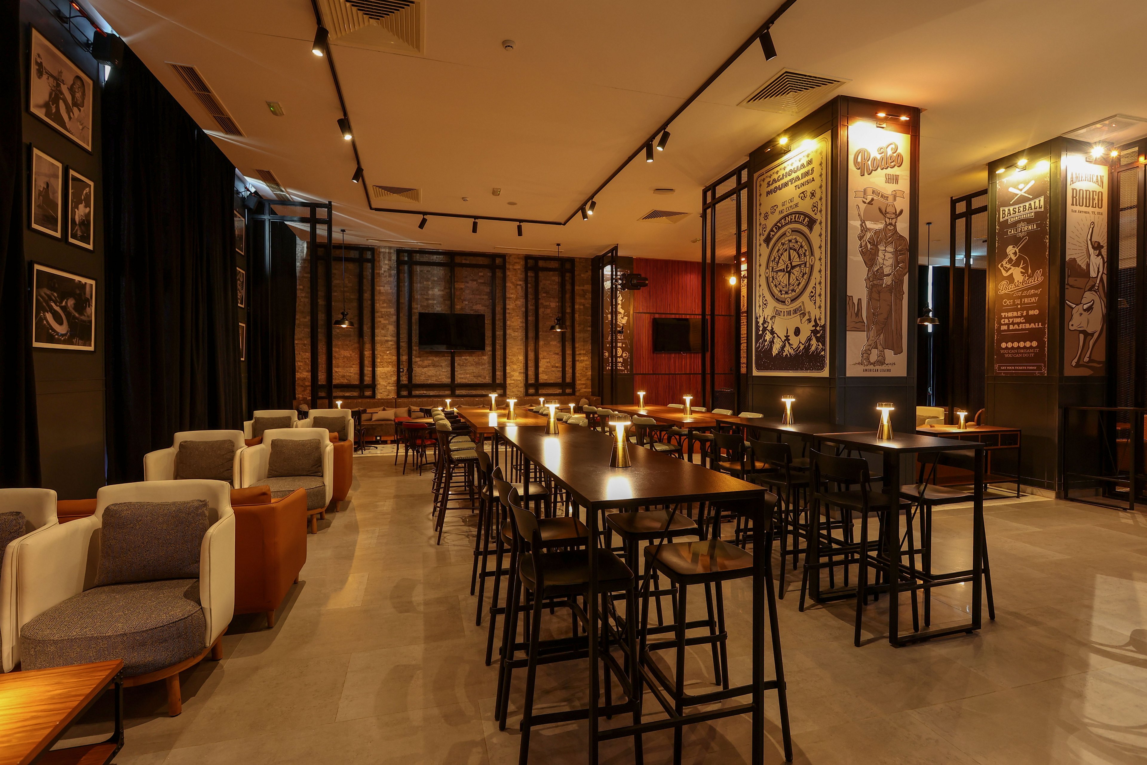 A modern bar interior features long high tables and stools arranged in neat rows under warm ambient lighting. Small table lamps create a cozy glow across the polished floor and dark ceiling. Large illustrated wall panels and framed artwork decorate the walls, adding visual interest and a contemporary feel. The space appears empty of people, emphasizing the inviting atmosphere and stylish design.