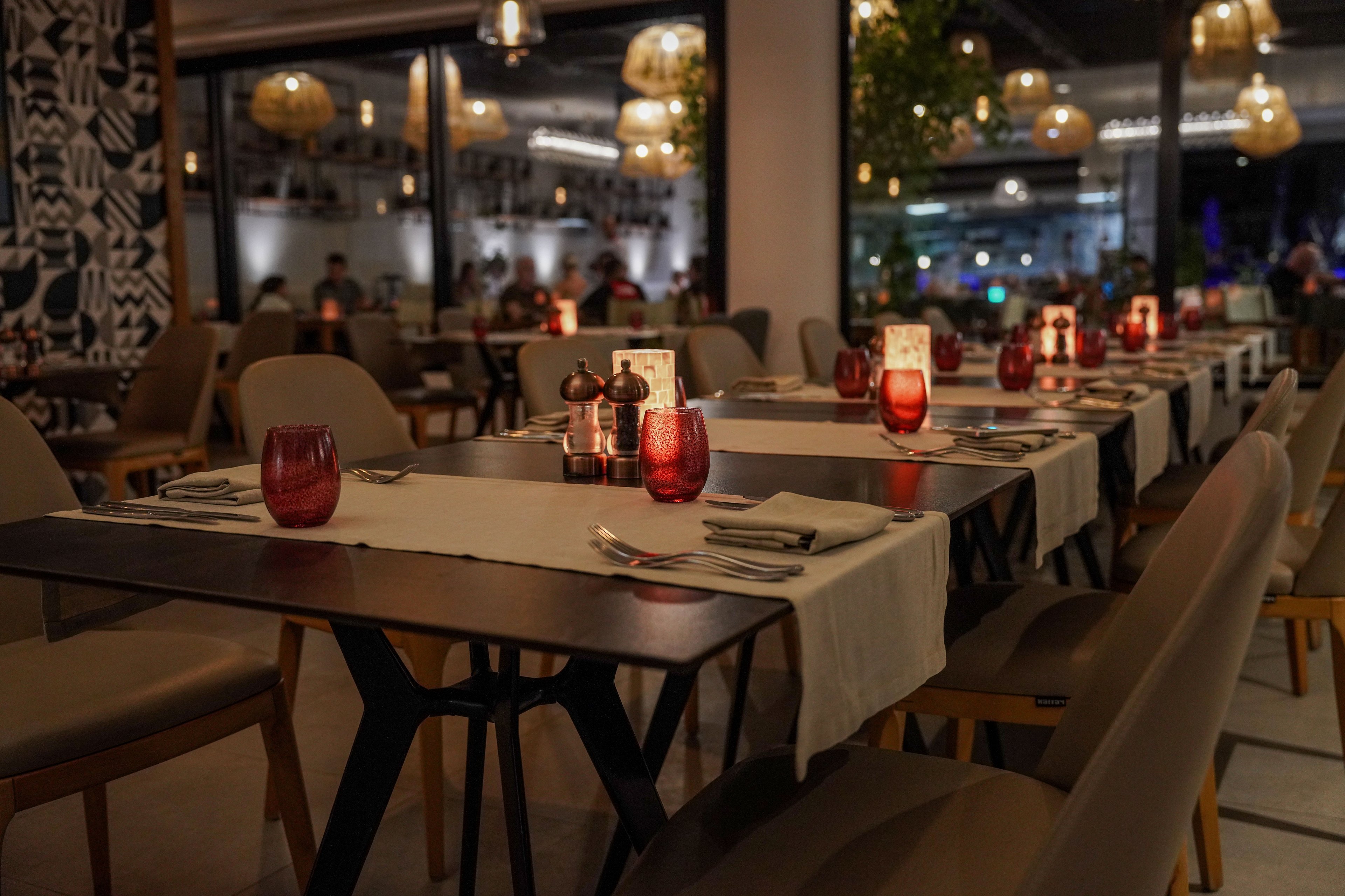 A warmly lit restaurant interior features neatly arranged dining tables set for guests. Each table is decorated with red glassware, candles, and folded napkins, creating an inviting atmosphere. Soft ambient lighting, hanging lamps, and modern chairs contribute to a cozy, upscale feel. The background shows additional seating and decor, suggesting a stylish evening dining experience.