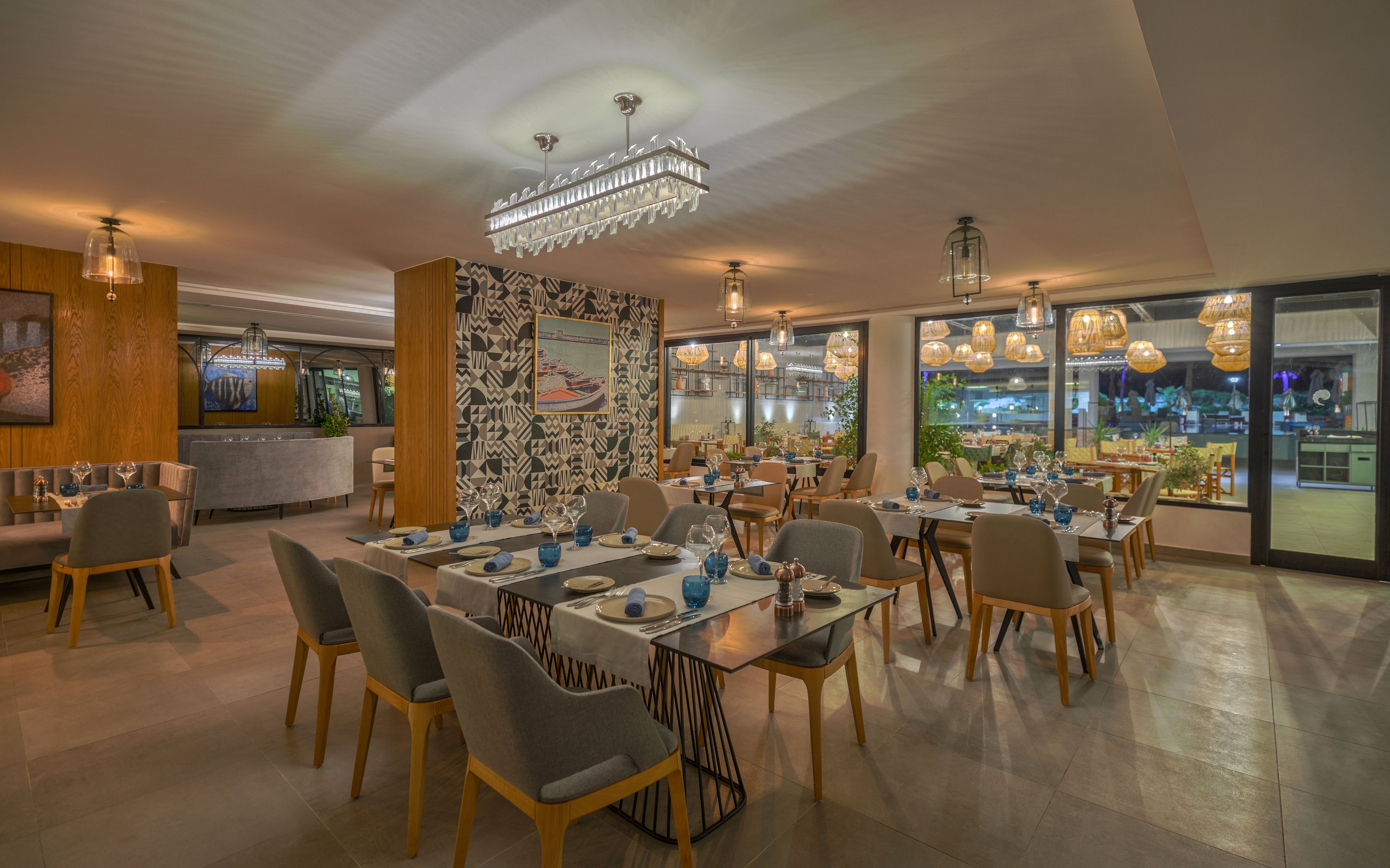 Contemporary restaurant interior featuring neatly set dining tables with blue glassware and neutral upholstered chairs. The space is warmly lit by chandeliers and pendant lights, creating a cozy evening atmosphere. Large windows reveal an outdoor area with greenery and additional seating. Decorative wall art and patterned partitions add visual interest to the modern design.