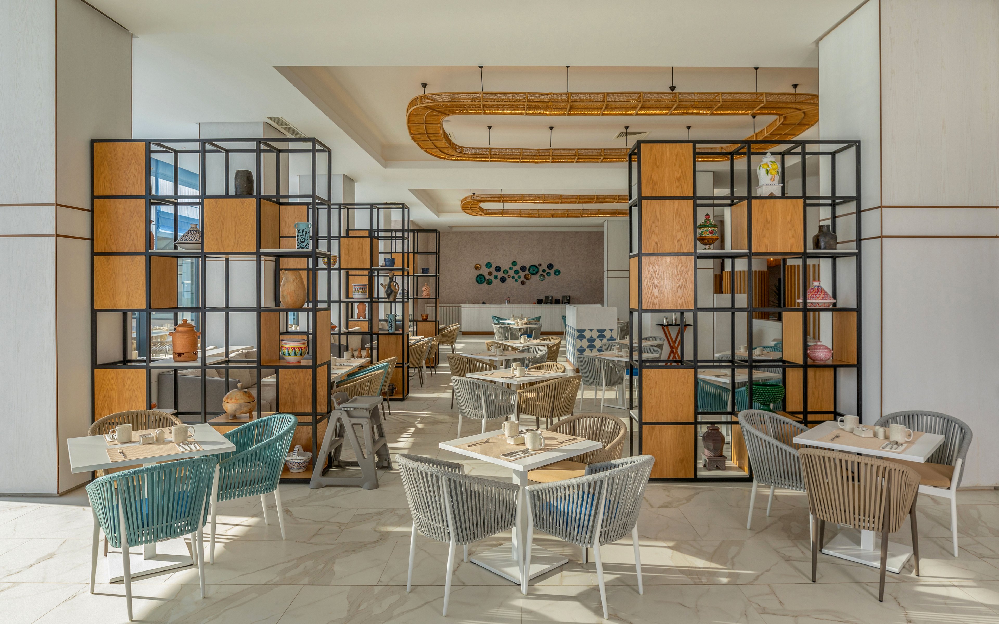 Contemporary restaurant seating is arranged in a bright, open hotel interior. Light-colored tables and woven chairs sit on a polished stone floor, framed by decorative shelving partitions with wood and metal accents. Natural daylight streams in from large windows, highlighting minimalist decor and sculptural elements on the shelves. The overall atmosphere feels relaxed, stylish, and welcoming for casual dining.
