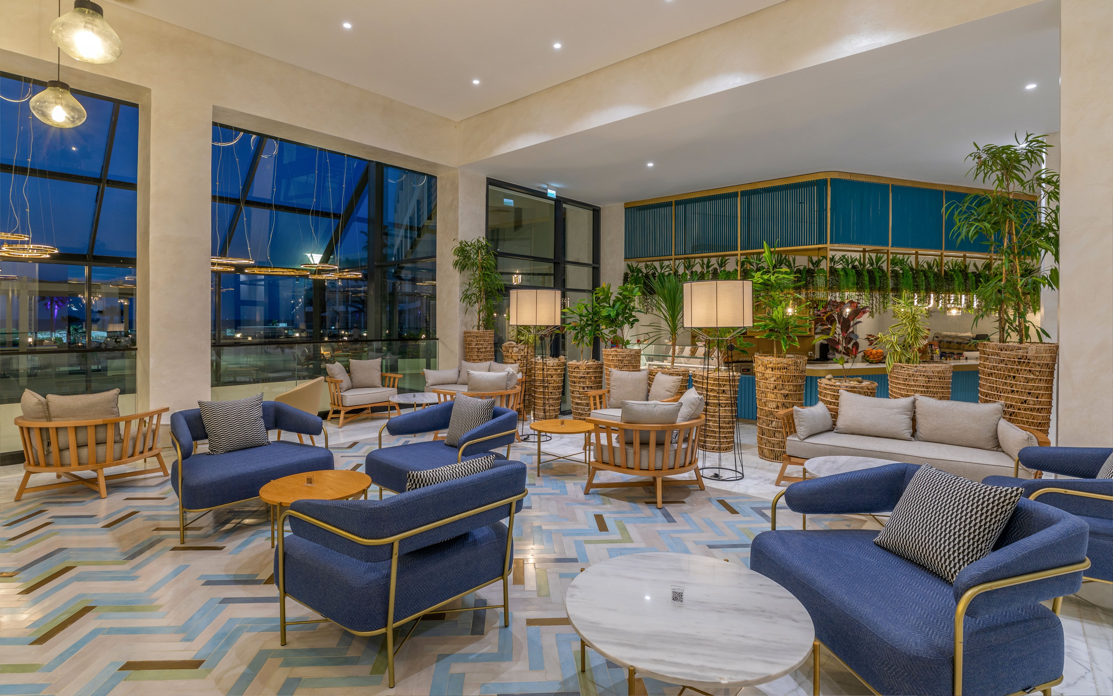 A contemporary hotel lounge features blue upholstered armchairs and light wooden furniture arranged in cozy seating groups. Large floor-to-ceiling windows reveal an evening city or waterfront view outside. The interior includes patterned geometric flooring, potted plants, and warm pendant and floor lighting that create an inviting atmosphere. A bar or café area with green accents and wicker details is visible in the background, enhancing the relaxed social setting.
