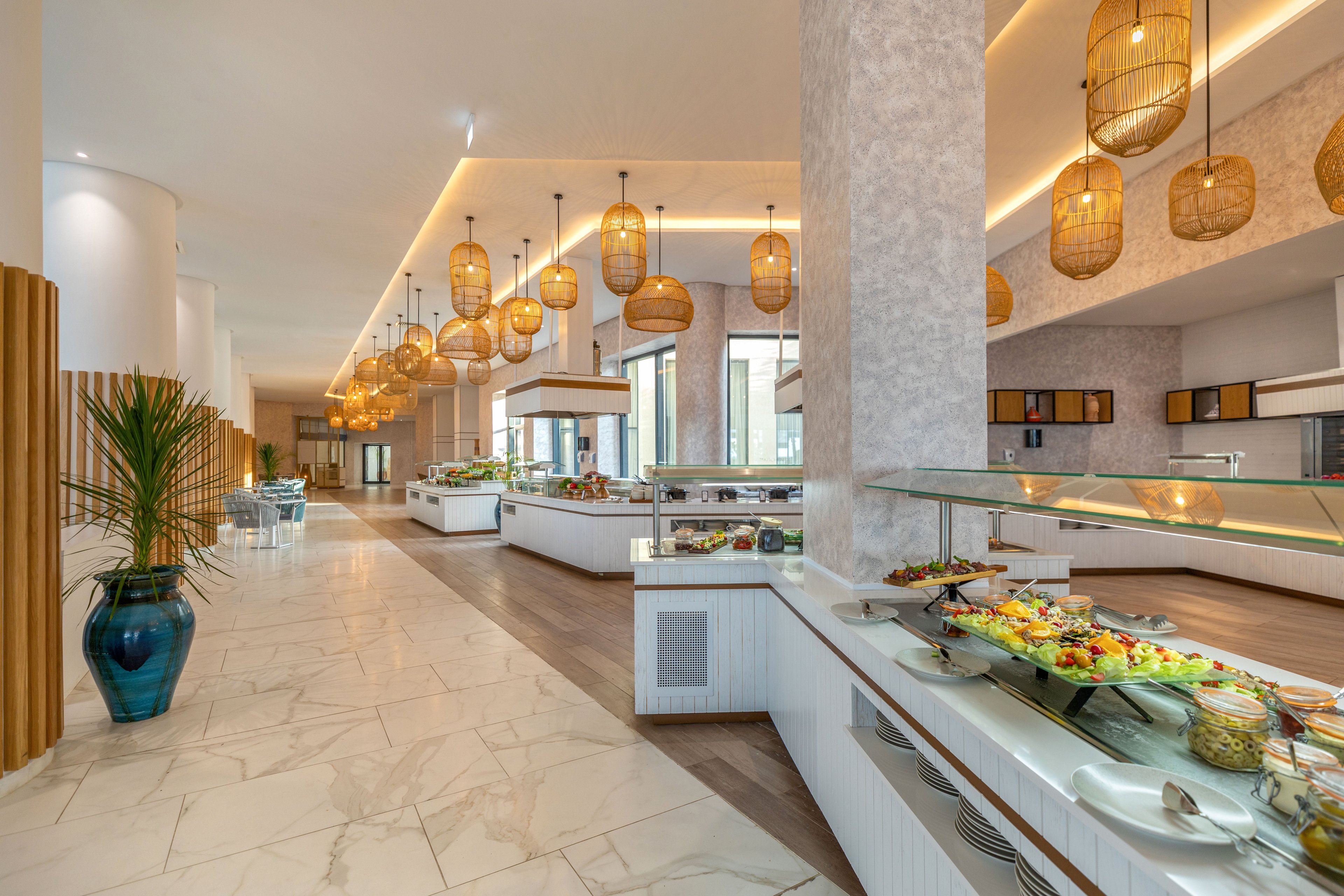 Bright, contemporary buffet restaurant interior with multiple food stations. A long counter in the foreground displays assorted salads, cold dishes, and bread under a glass sneeze guard. Warm pendant basket lights hang from the ceiling, complementing the light marble flooring and neutral walls. Large windows and decorative plants add an airy, inviting atmosphere to the spacious dining area.